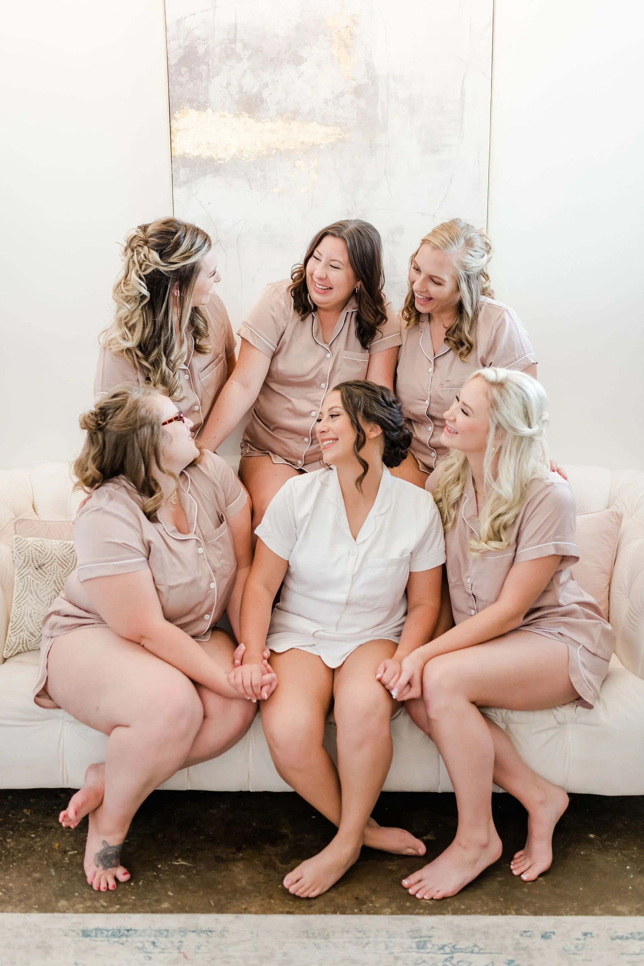 Seven women in coordinated pajamas sitting and standing on a cream couch, smiling and holding hands, celebrating a wedding or special occasion.