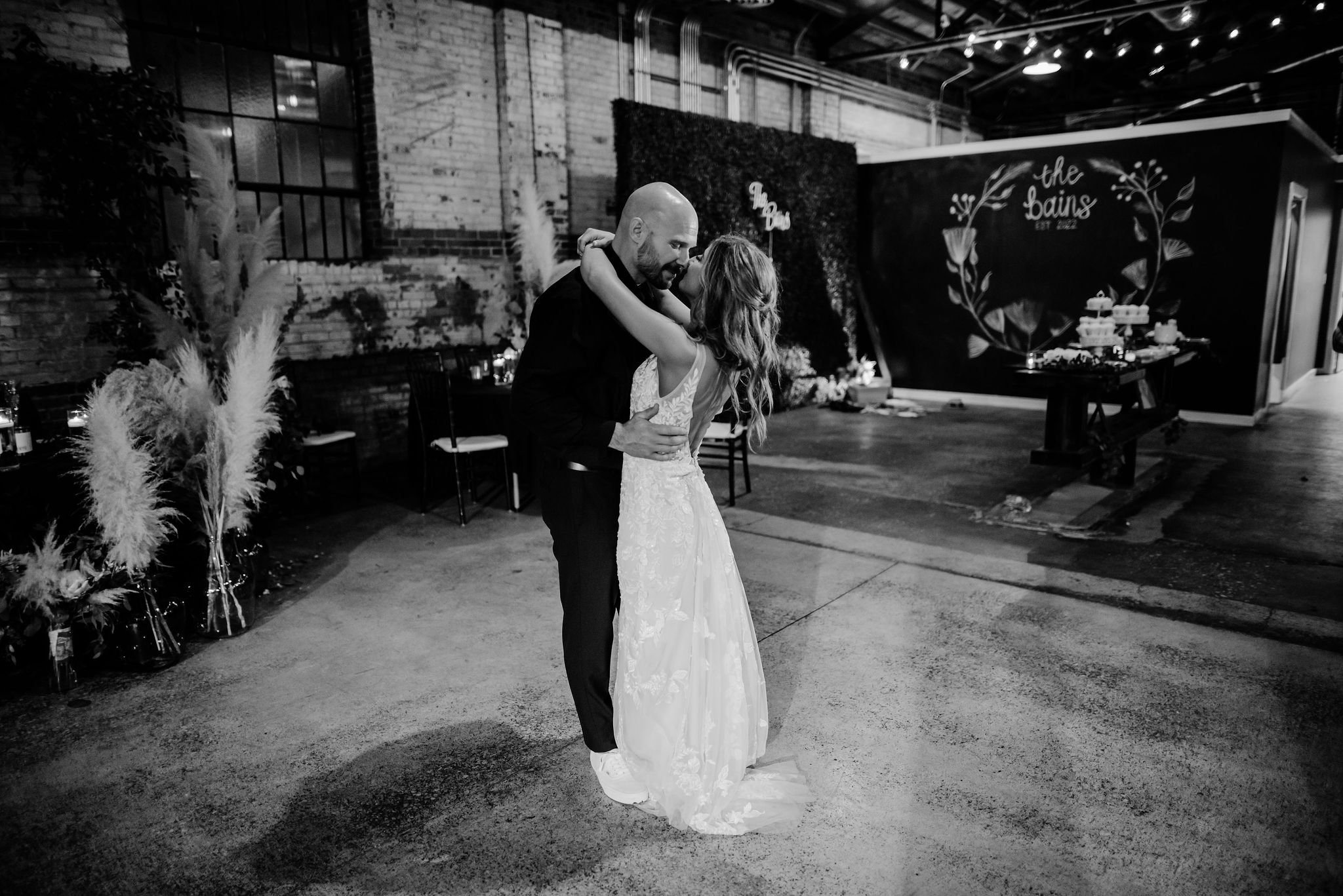 A couple dancing at their wedding reception. The woman is wearing a white lace wedding dress, and the man is dressed in a dark suit. They are embracing in a decorated indoor venue with a backdrop reading "The Bains."