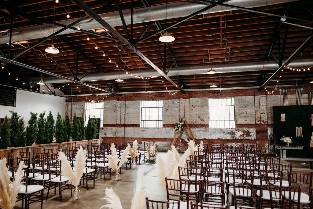 Interior of a rustic industrial-style event space with rows of chairs, boho decor, string lights, and greenery for a wedding or celebration.