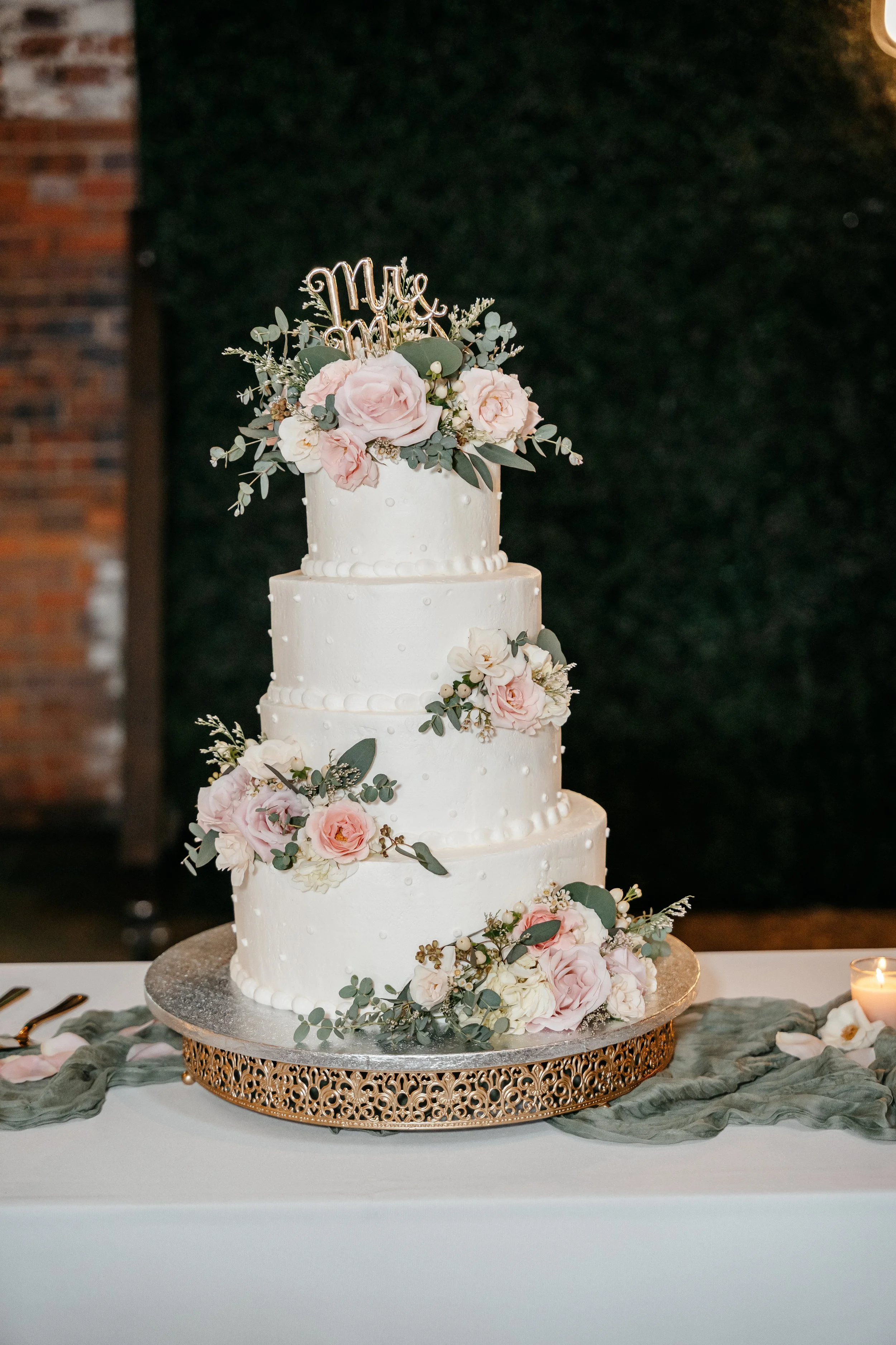 A four-tiered white wedding cake decorated with pink roses, white flowers, and greenery, topped with a "Mrs & Mr" cake topper, on a decorated table with a candle and pink flower petals.