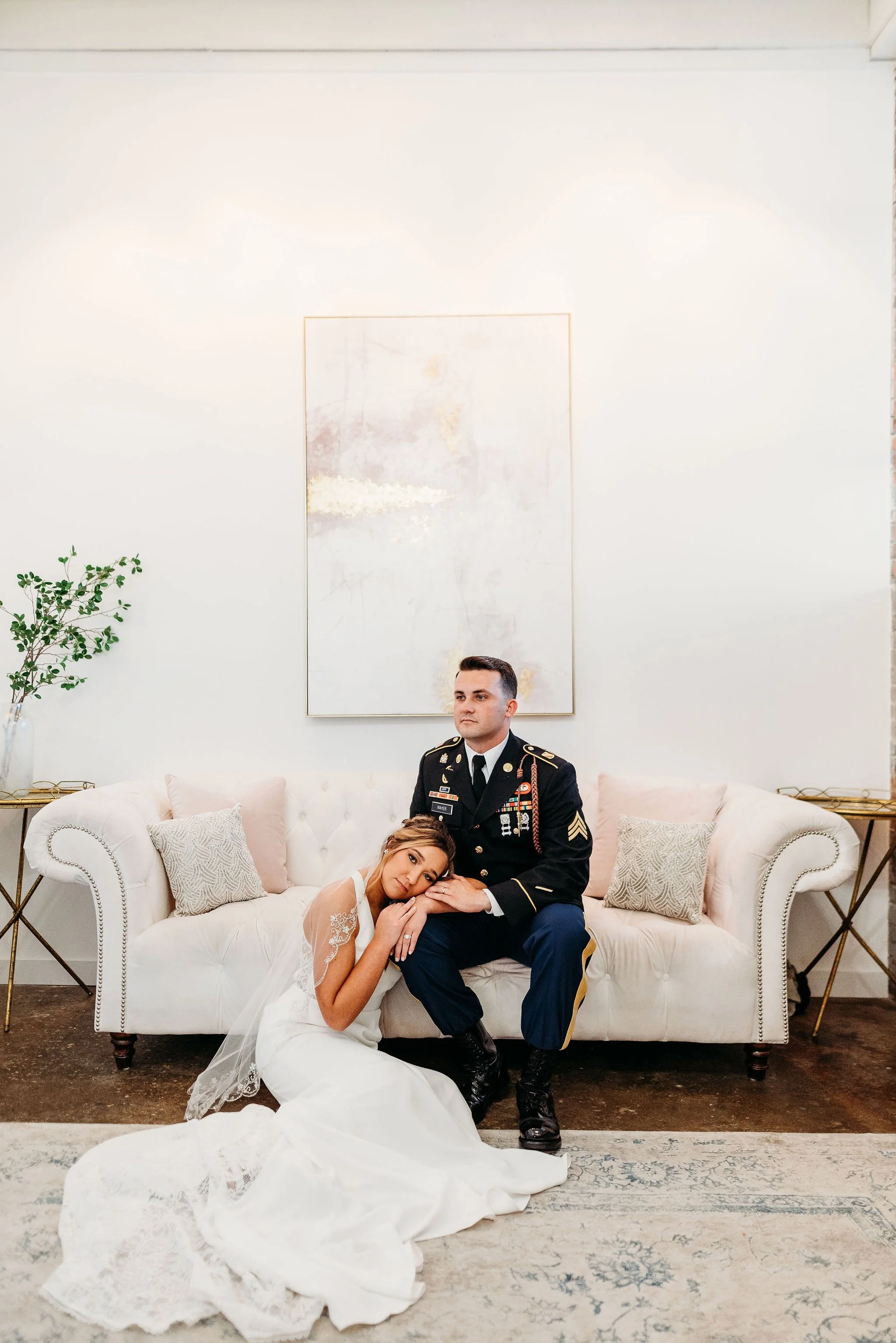 A bride leaning her head on the lap of a groom in a military uniform, sitting on a white sofa in a modern, minimalist room.