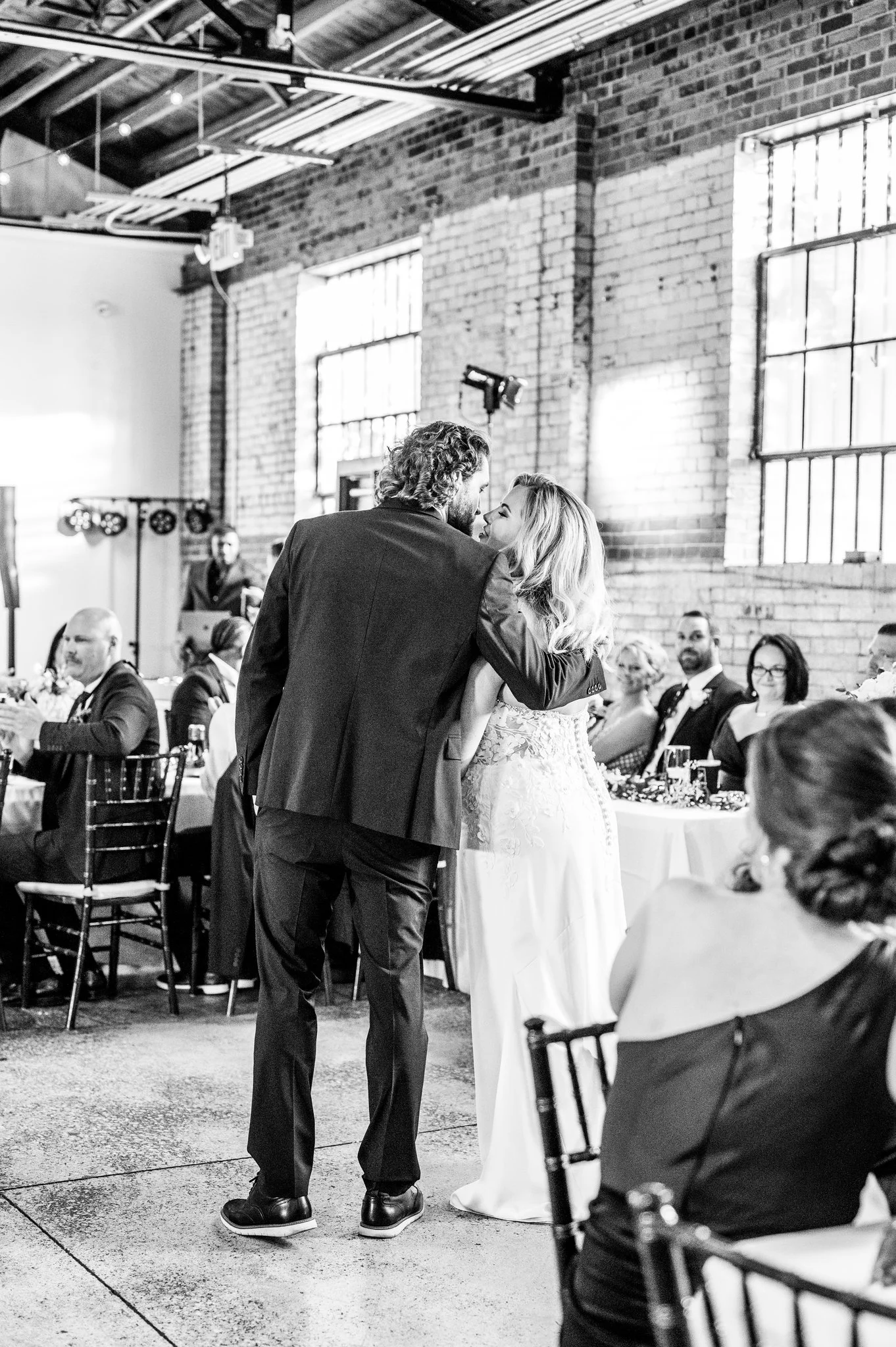Black and white photo of a wedding reception showing a bride and groom sharing a dance, surrounded by seated guests in an industrial-style venue with brick walls and large windows.