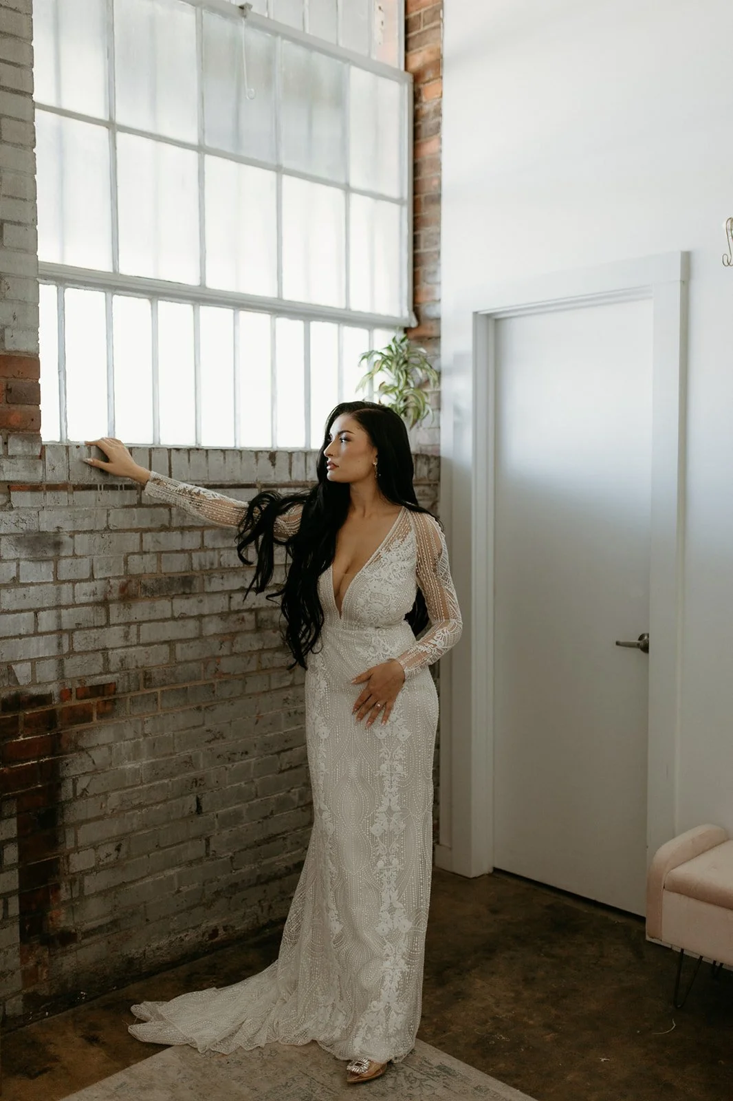 A woman in a long, white, lace wedding dress with long sleeves and a deep V neckline, standing indoors near a brick wall and a large window, with a potted plant nearby.