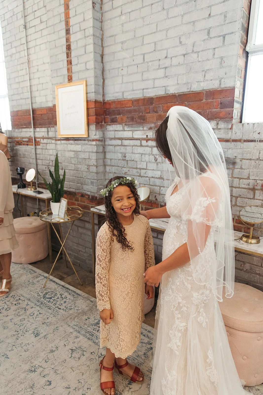 A bride in a lace wedding dress and veil smiling at a young girl with a floral crown and a beige lace dress, in a room with brick walls and gold-accented furniture.
