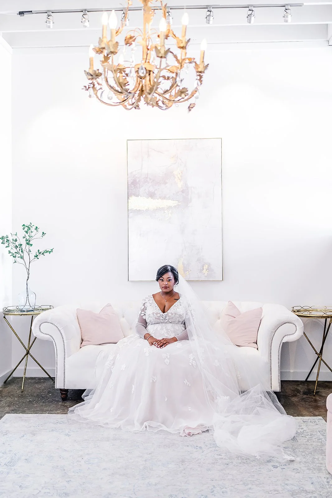 A bride in a white wedding dress sitting on a white sofa with pink pillows in a bright, minimalist room. There is a chandelier hanging from the ceiling, a large abstract painting on the wall behind her, and small side tables with decorative plants on each side of the sofa.