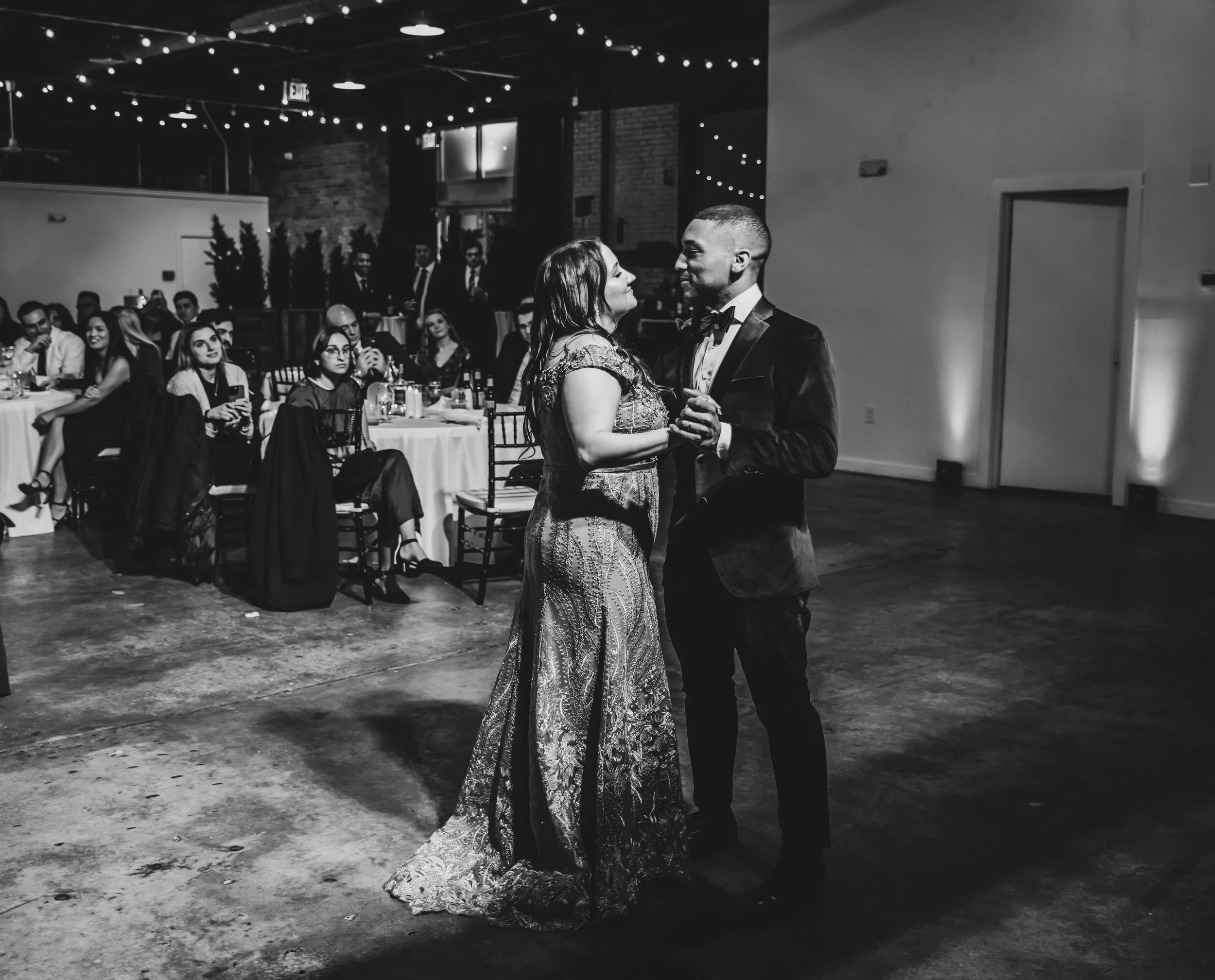 A couple dances together at a formal event with guests seated at tables in the background. The scene is indoors, decorated with string lights.