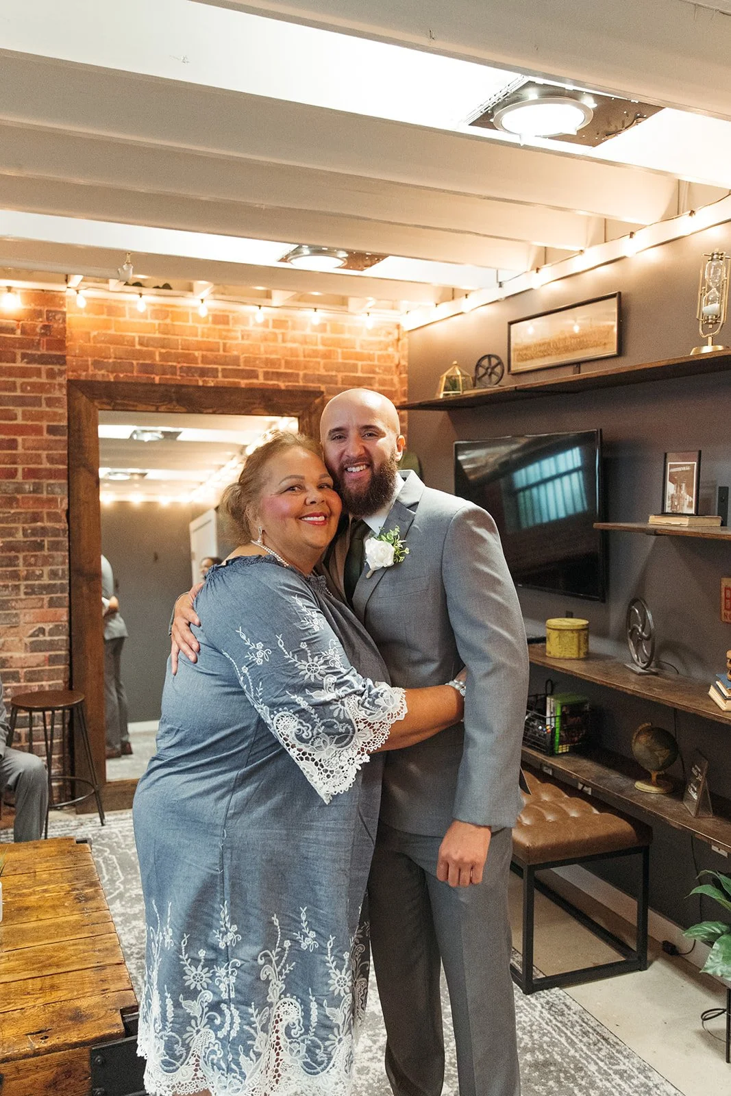 A woman in a gray dress hugging a man in a gray suit and black tie, both smiling, in an indoor setting with a brick wall and shelves in the background.