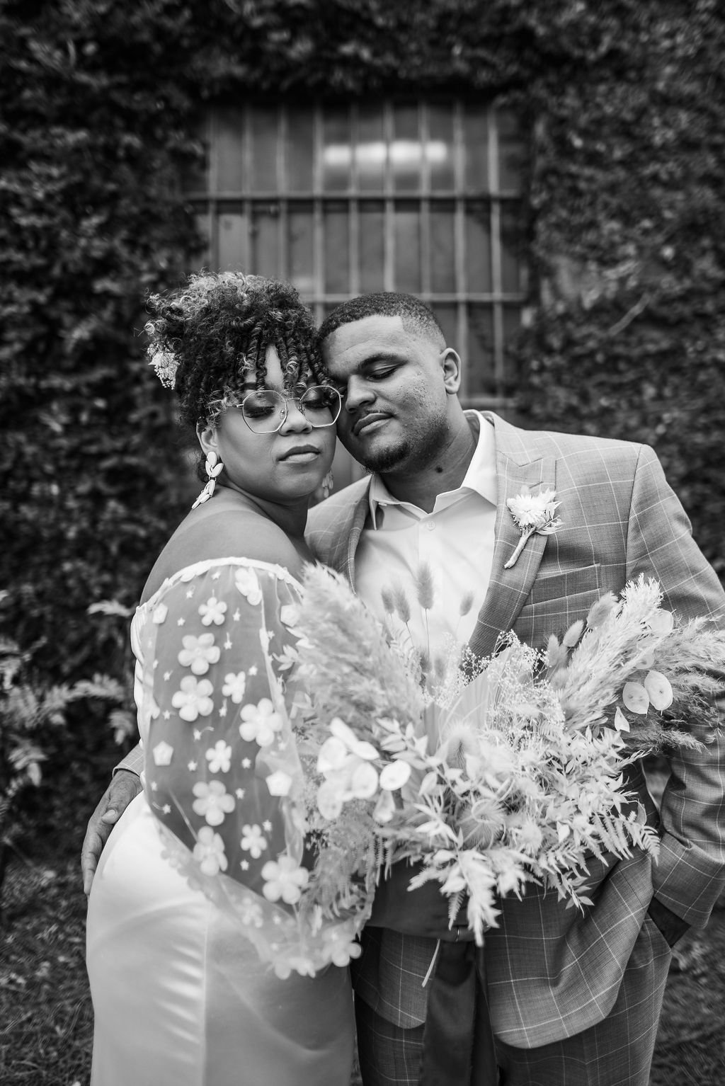 A black and white photo of a couple, with the woman wearing glasses and a floral dress, and the man in a checkered suit with a boutonniere, holding a large bouquet; they are close with their foreheads touching, in front of a brick wall with a window.