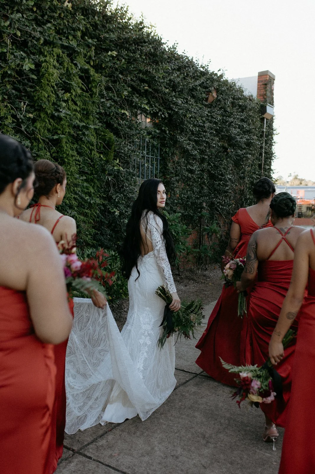 A bride in a white lace wedding dress holding a bouquet standing outdoors, surrounded by bridesmaids wearing red dresses and holding flowers, with a green ivy-covered wall in the background.