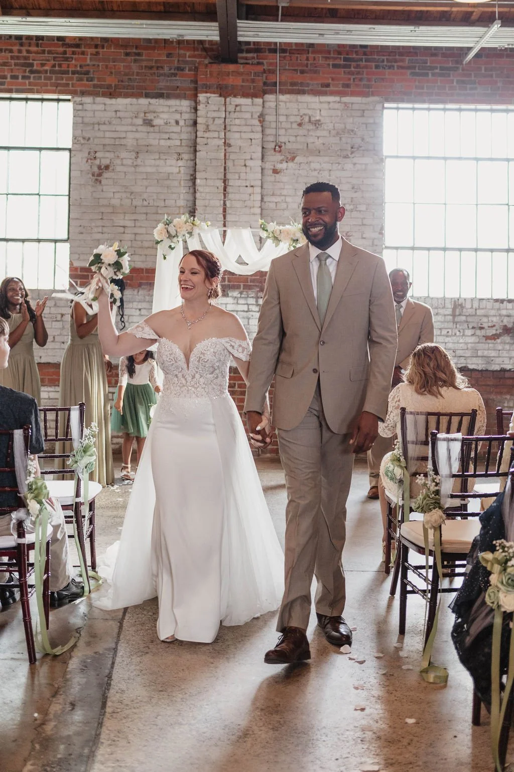 A newlywed couple walking hand-in-hand down the aisle of a wedding ceremony, smiling and celebrating with friends and family in a rustic indoor venue decorated with flowers and white drapery.