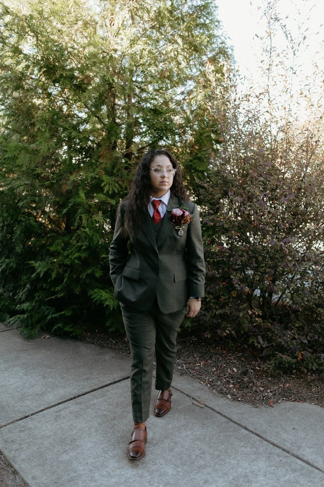 A woman in a dark gray suit with a red tie and a flower boutonniere, walking on a sidewalk outdoors with green trees and shrubs in the background during daytime.