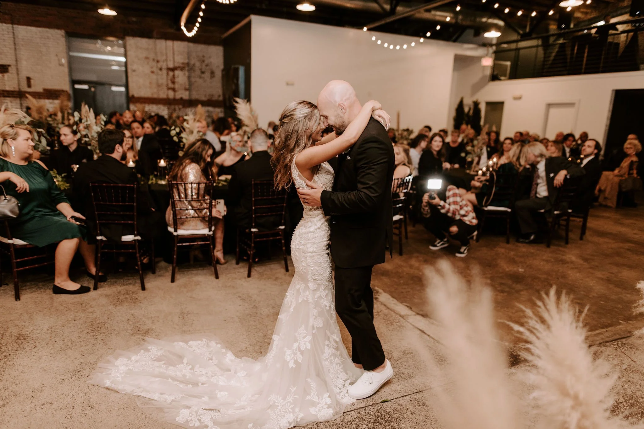 A bride and groom sharing a dance in front of seated guests at a wedding reception, with warm ambient lighting and rustic decor.