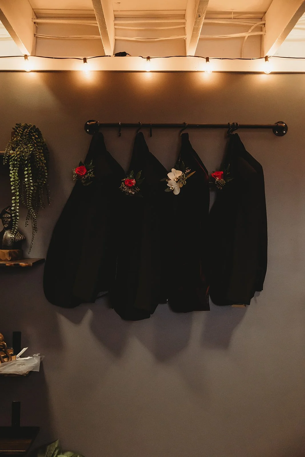 Four black suits or dresses hanging on a wall-mounted rod, decorated with flowers featuring red roses and white blossoms, with string lights above. A potted plant and a small bronze bust are visible on a shelf to the left.
