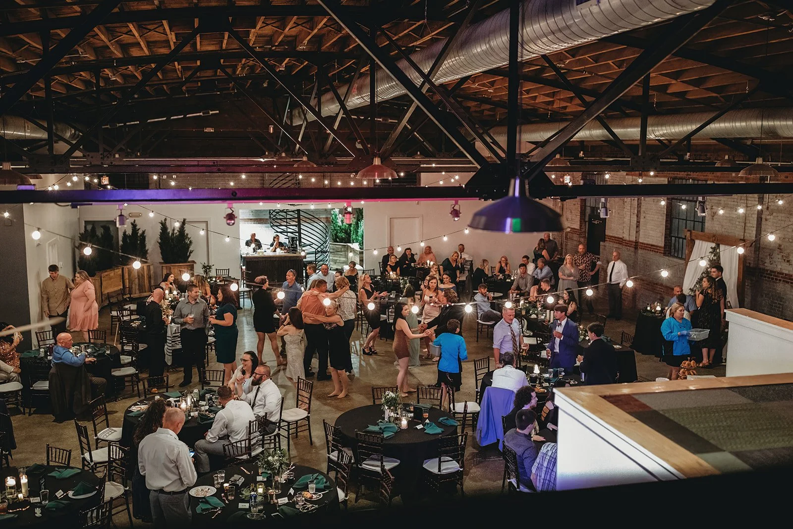 A lively indoor party or wedding reception with guests socializing, dancing, and sitting at decorated tables in a spacious, industrial-style venue with string lights and brick walls.