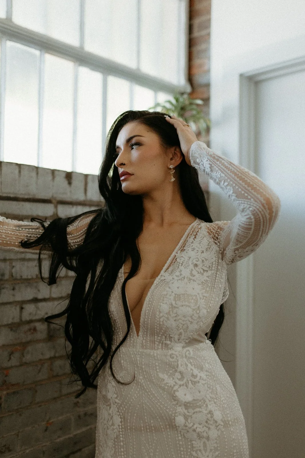 A woman with long dark hair wearing a white lace dress, posing with one hand on her head in a well-lit room with large windows and a brick wall in the background.