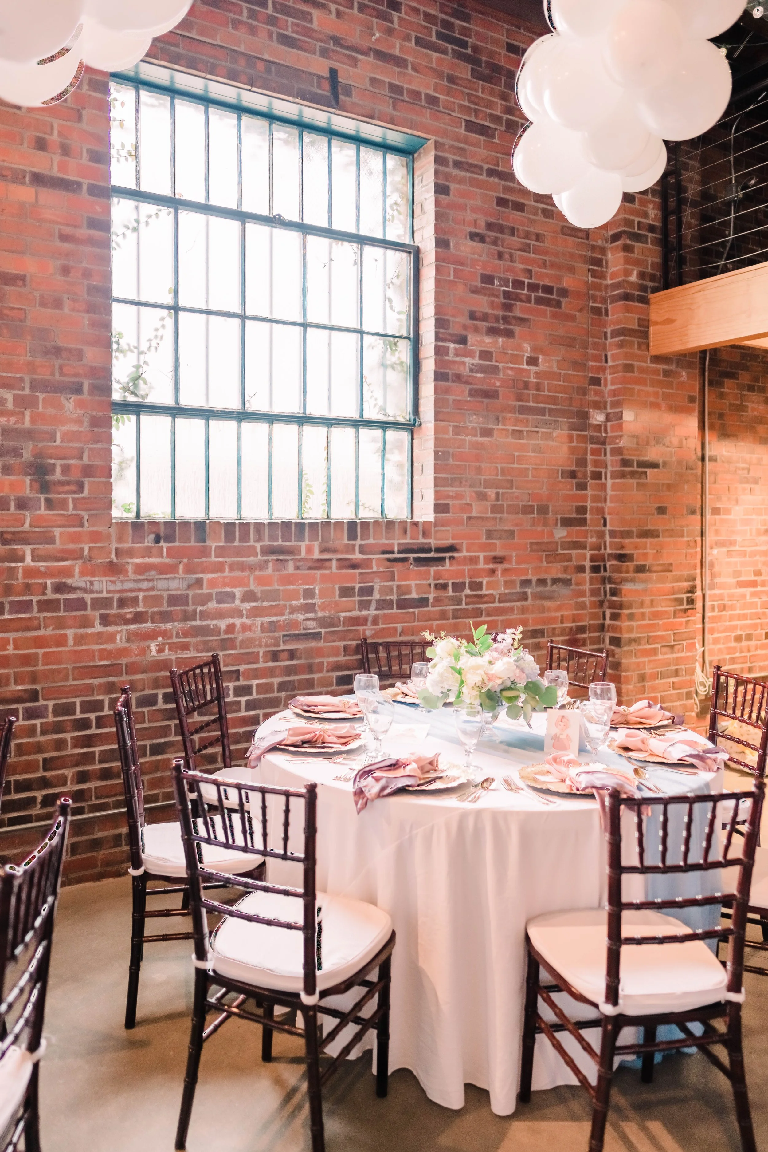 Round table set for a celebration with elegant tableware, pink napkins, and a floral centerpiece in a room with exposed brick walls, large window, and white balloons hanging from the ceiling.