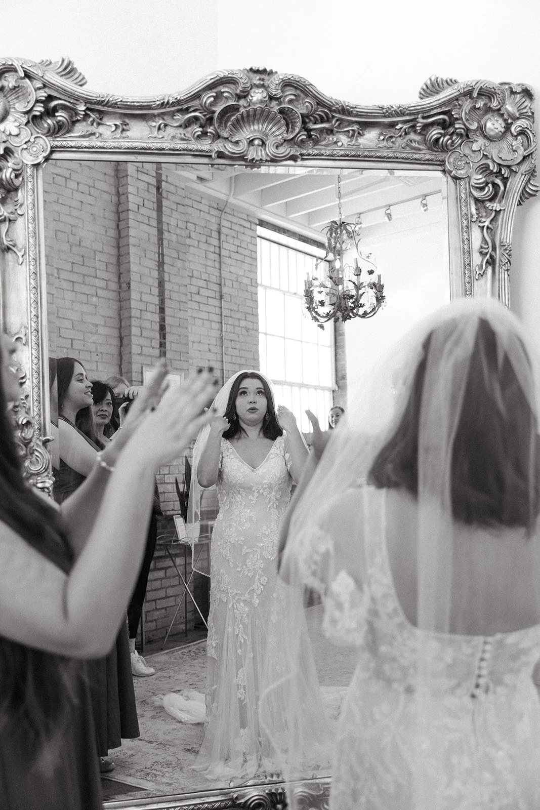 A bride putting on earrings in front of a large ornate mirror, with bridesmaids and friends watching, in a wedding dress and veil, in a room with brick walls and large windows.