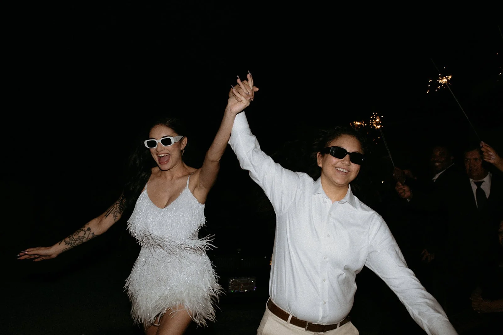 Two women wearing sunglasses and smiling, celebrating with sparklers at night.