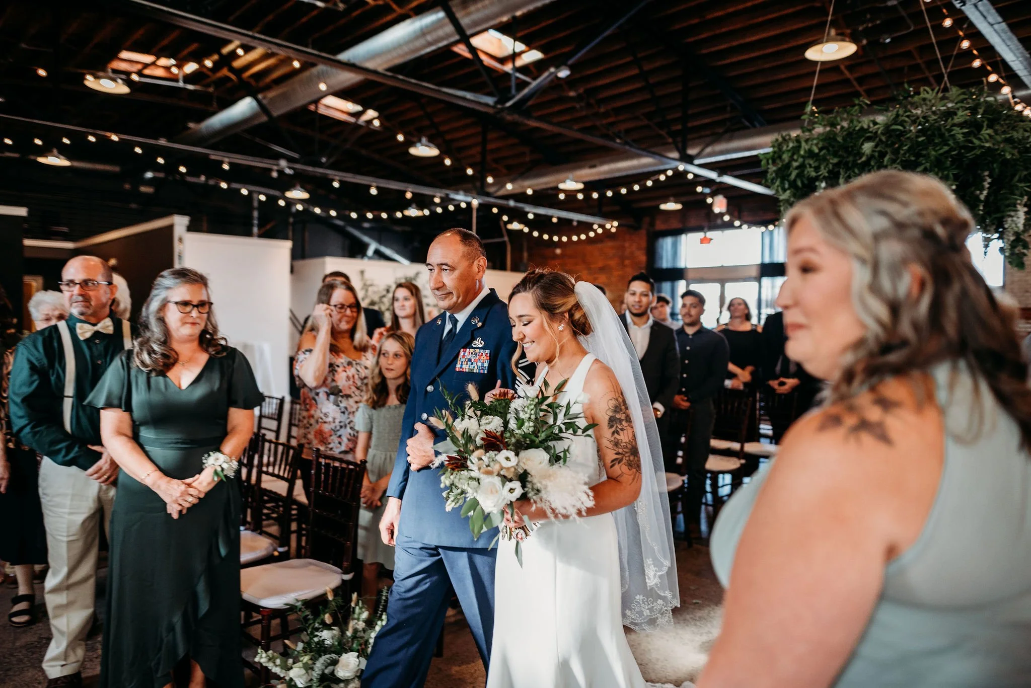 Bride walking down the aisle with her father at her wedding ceremony, surrounded by guests inside a venue with string lights and an industrial-style ceiling.