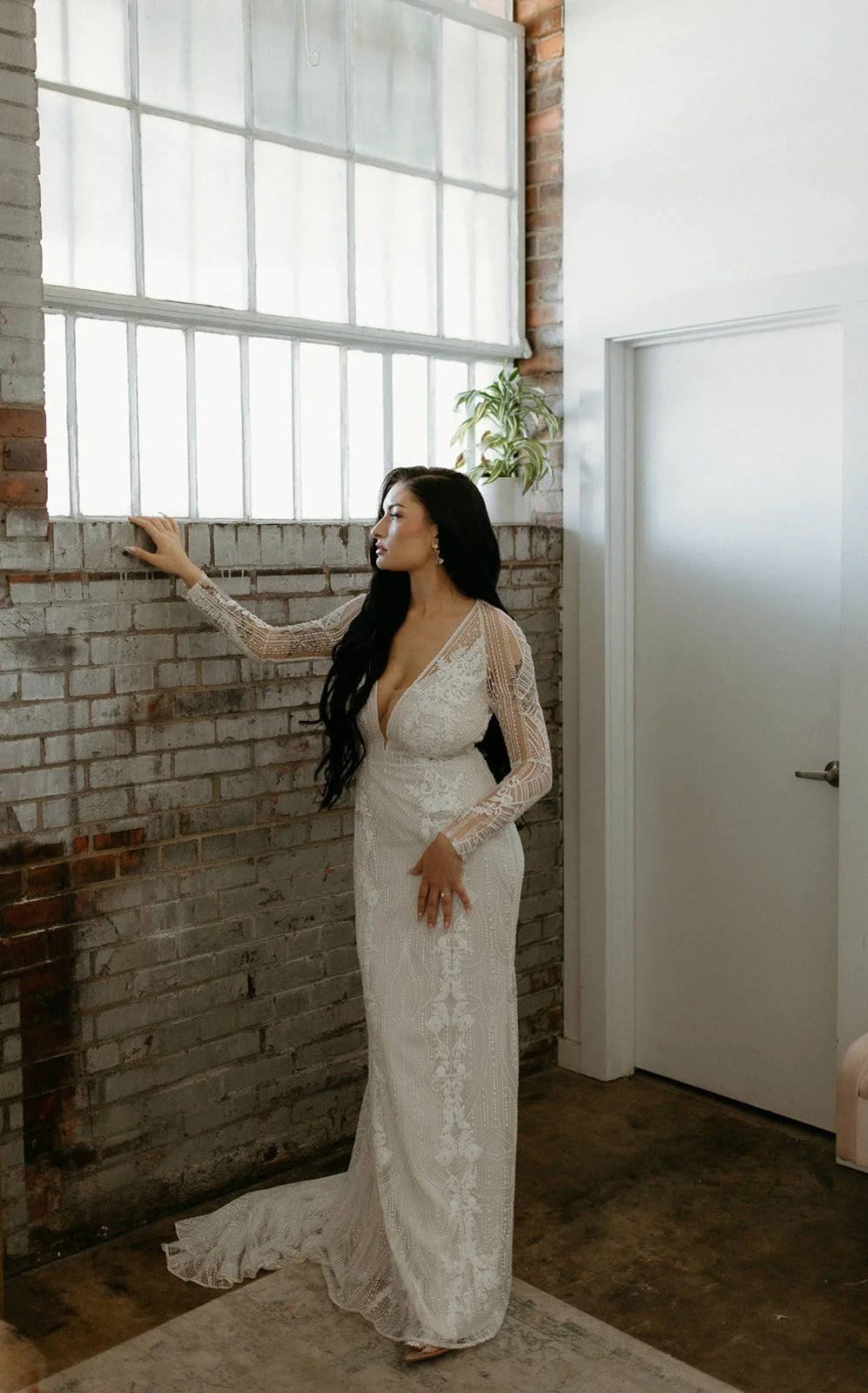 Elegant bride in long-sleeve lace wedding dress posing against an industrial exposed brick wall at Studio 215, a premier wedding venue in Fayetteville, NC