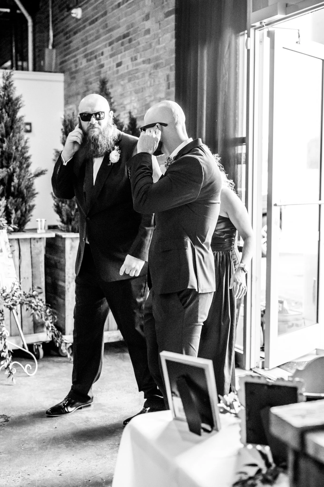 Two men in formal suits and sunglasses talking at an event, with a woman in a dress in the background near an open door.