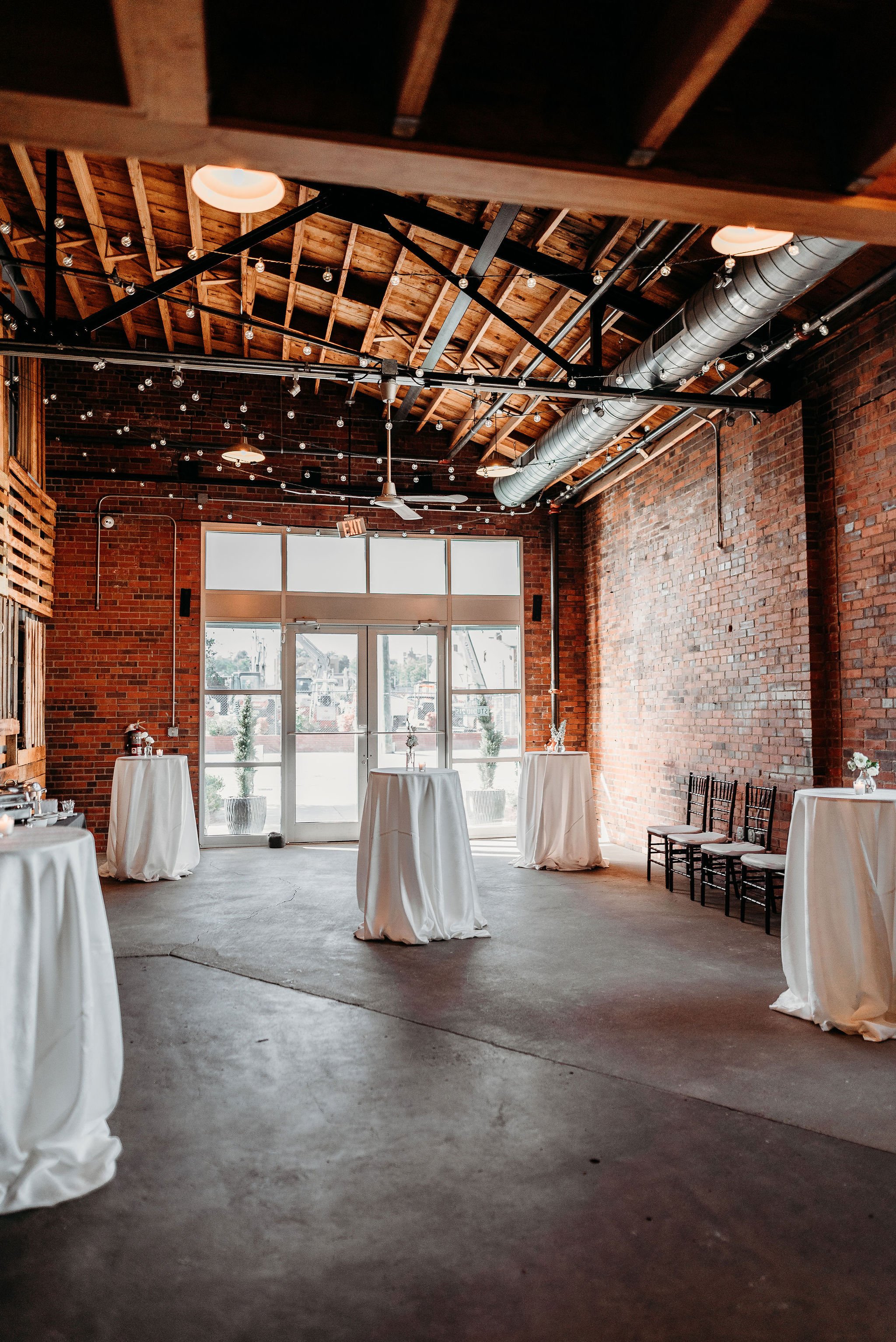Empty brick-walled event space with high ceiling, string lights, and tables draped in white cloth, with a glass double door at the back.