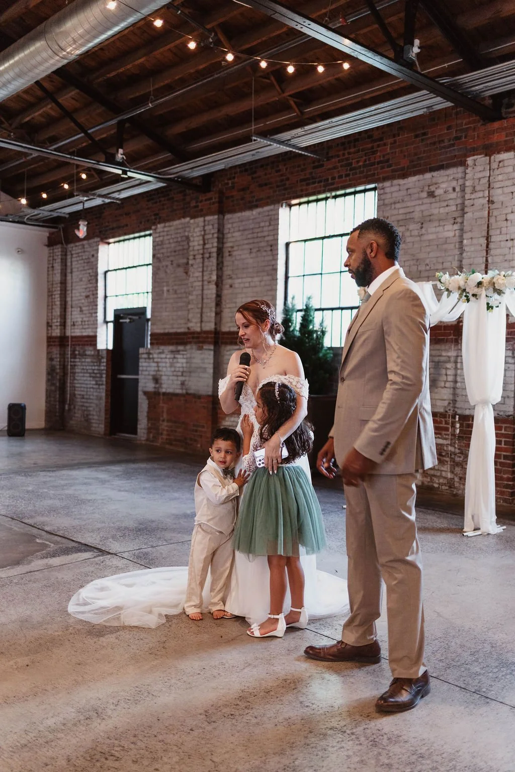 A bride in a white wedding dress holding a microphone, standing next to a groom in a beige suit, with two children in front of her during a wedding ceremony in an industrial-style venue with brick walls and large windows.