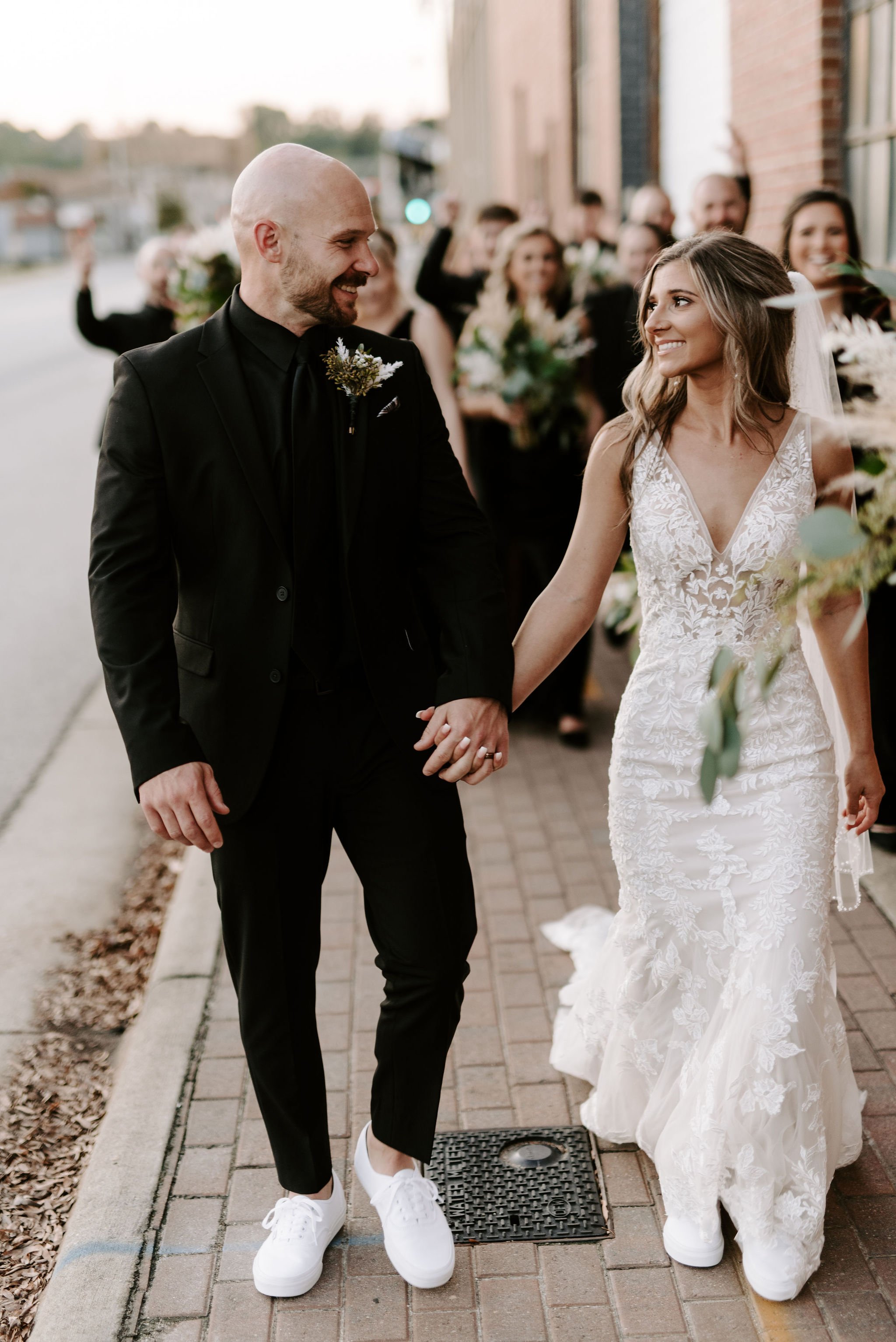 A bride and groom holding hands and smiling at each other during their wedding ceremony on a city sidewalk, with wedding guests in the background.