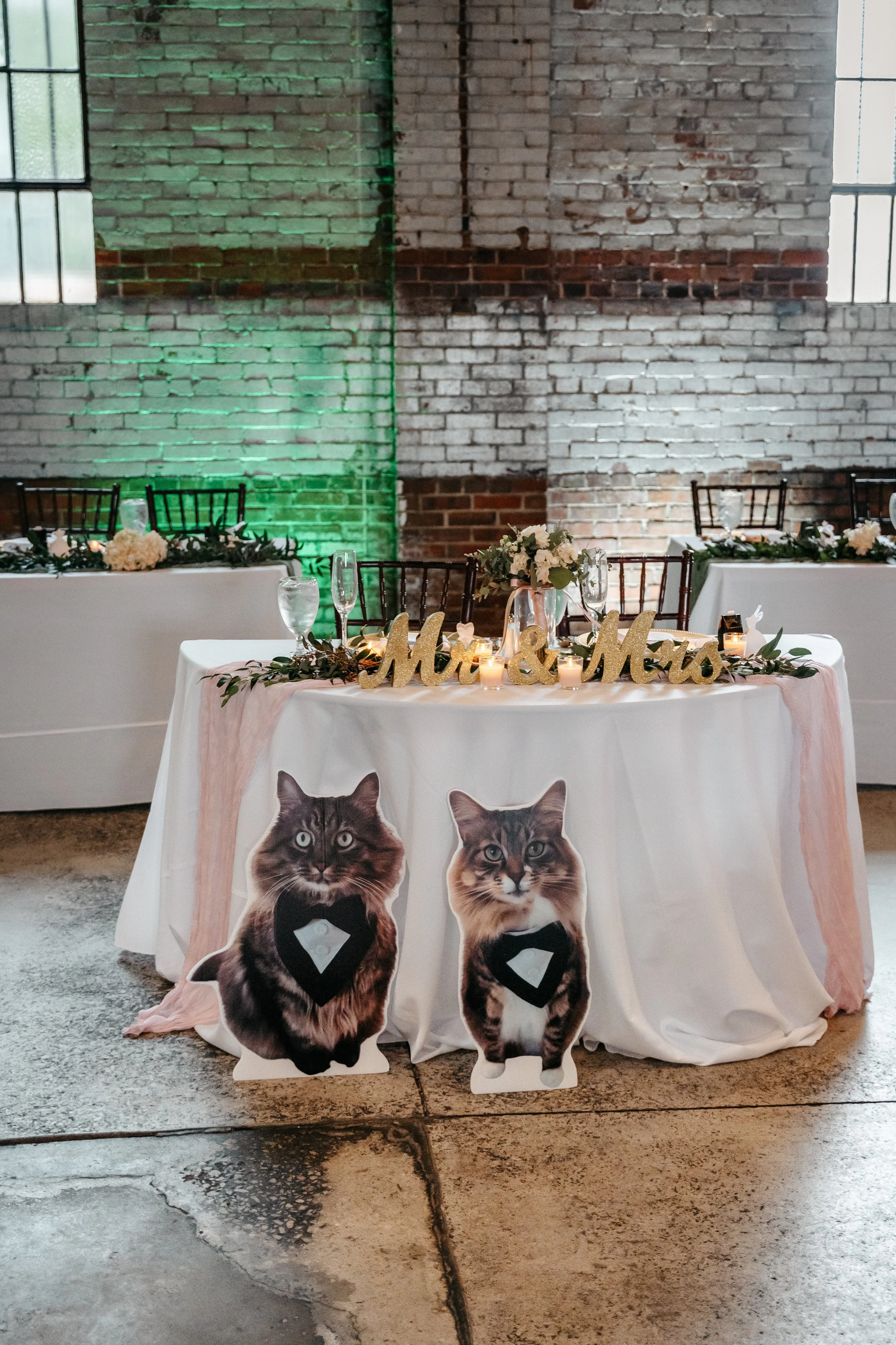 Decorated sweetheart table for a wedding with 'Mr. & Mrs.' sign, floral arrangements, candles, and cardboard cutouts of cats with bow ties in front.