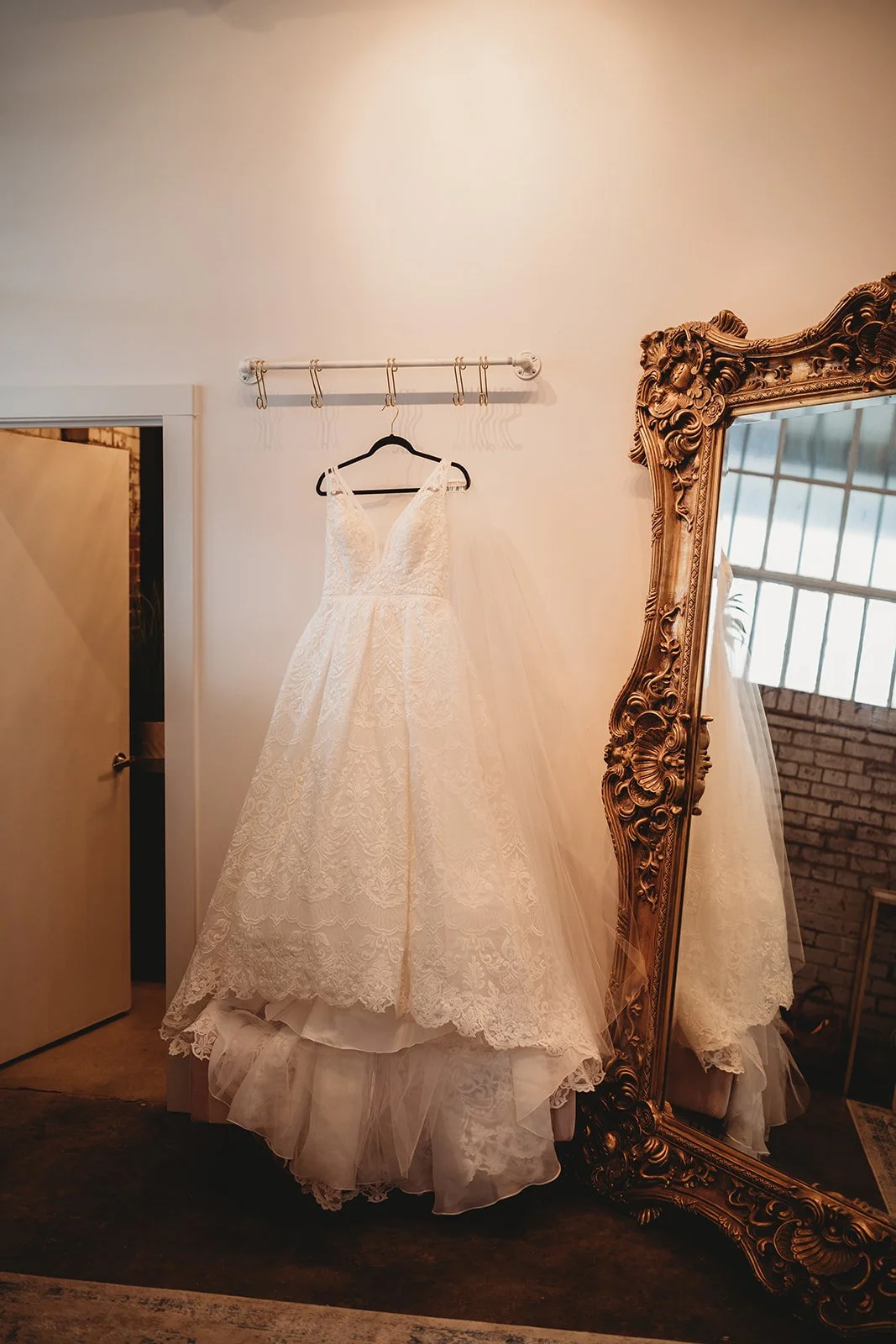 A white lace wedding dress hanging on a black hanger on a white wall, reflected in an ornate gold mirror.
