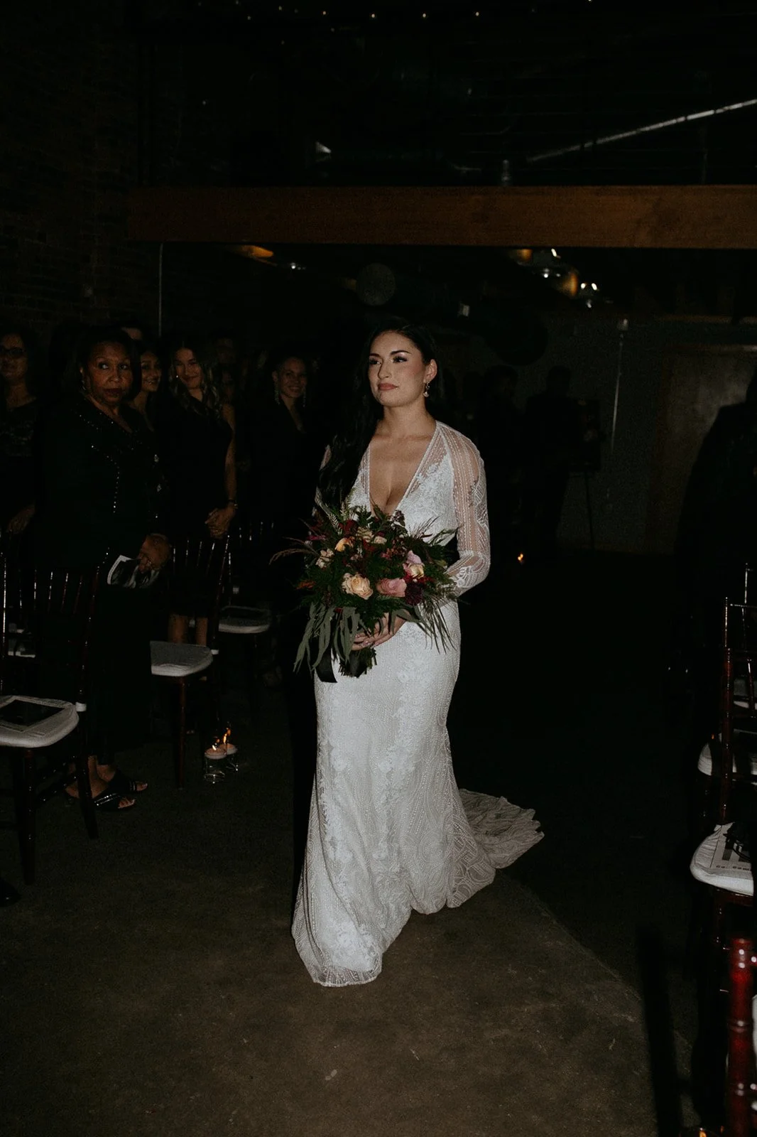 A bride in a white lace wedding dress holding a bouquet of pink and red flowers walking down the aisle in a dimly lit indoor venue, with several guests standing and seated in the background.