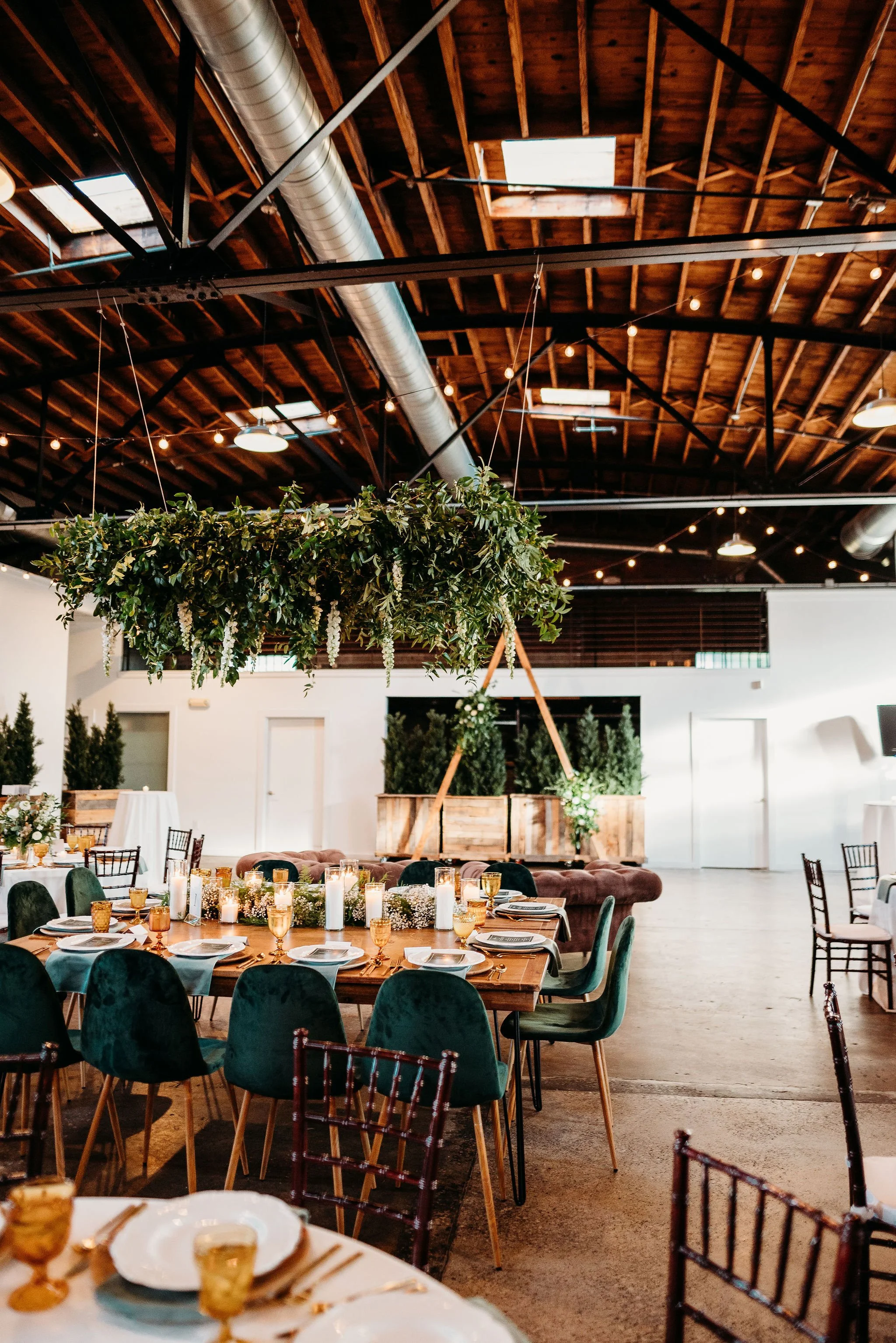 Event space decorated with a large circular wooden table with plates, glasses, and candles, hanging greenery centerpiece, string lights on a wooden ceiling, and potted plants in the background.