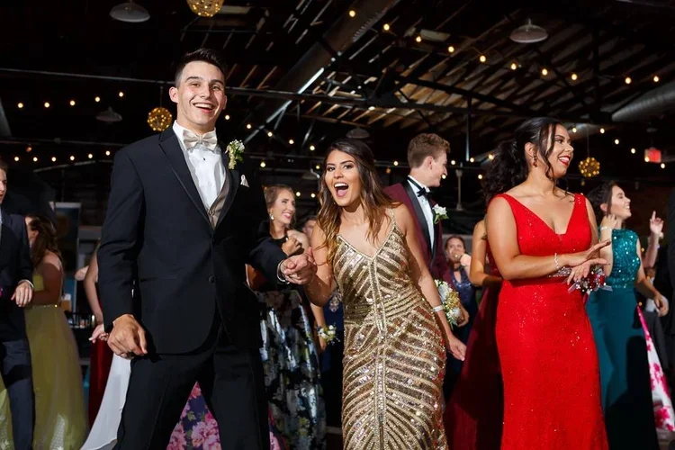 People dancing at a wedding reception, including a smiling man in a tuxedo and a woman in a gold dress, with other guests in formal attire under string lights.