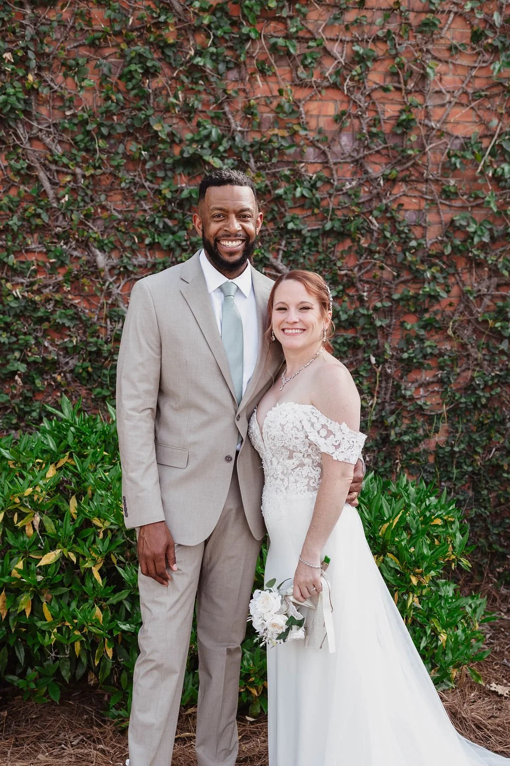 A happy interracial couple in wedding attire posing outdoors in front of a brick wall covered in green ivy. The groom wears a light gray suit with a white shirt and light blue tie, and the bride wears a white off-shoulder wedding gown with lace detai