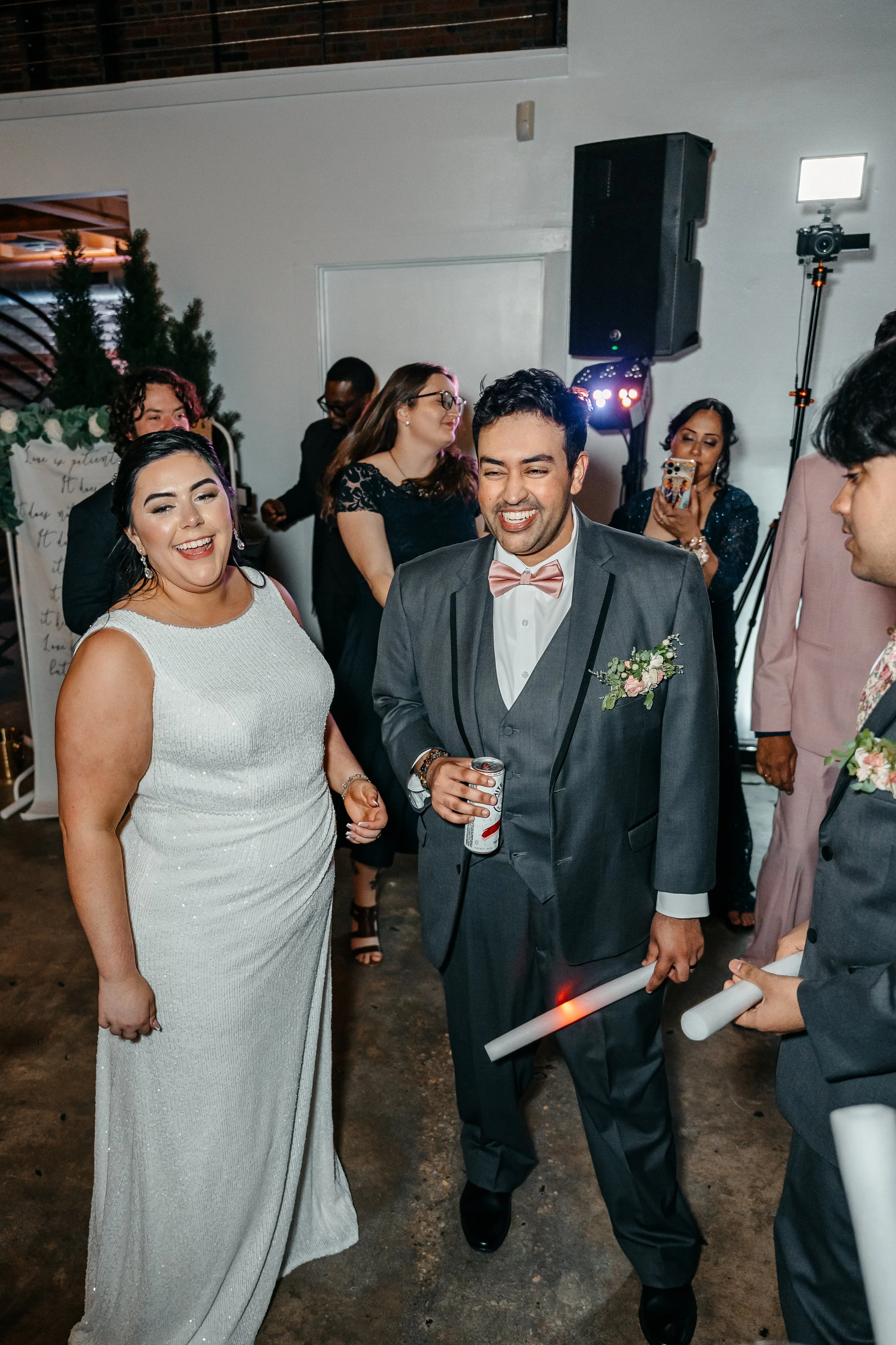 People dancing and celebrating at a wedding reception, with a smiling bride and a groom holding a drink, in a decorated indoor venue.