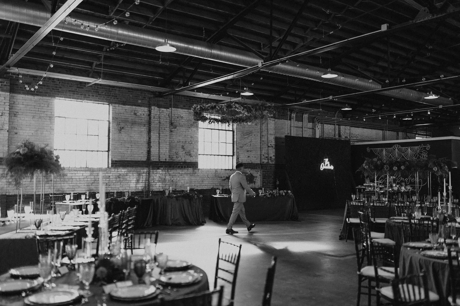 Black and white photo of an indoor event space with decorated tables, a man walking, and a large decorated wall with a sign. The space has exposed brick walls, large windows, and industrial ceiling structures.