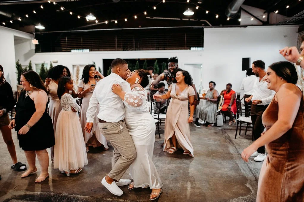 A diverse group of people dancing and celebrating indoors at a wedding reception, with a bride and groom dancing in the center.