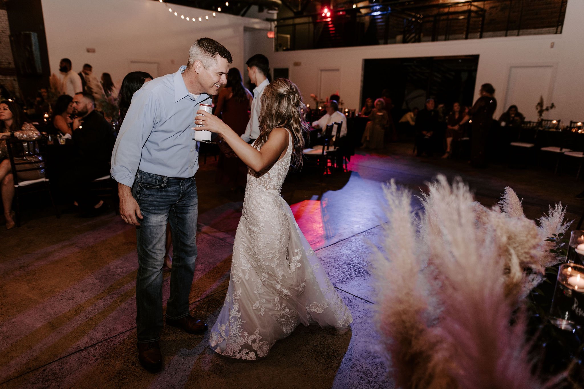 A bride and an older man dancing together at a wedding reception, with guests sitting at tables in the background.