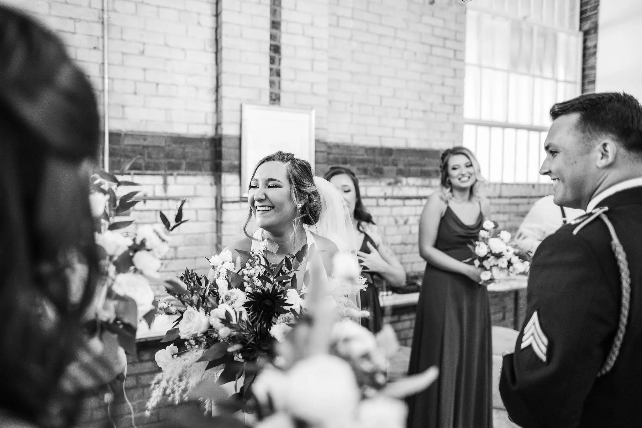 Black and white photo of a wedding ceremony with a bride smiling at the groom, surrounded by bridesmaids holding bouquets, with a brick wall background.