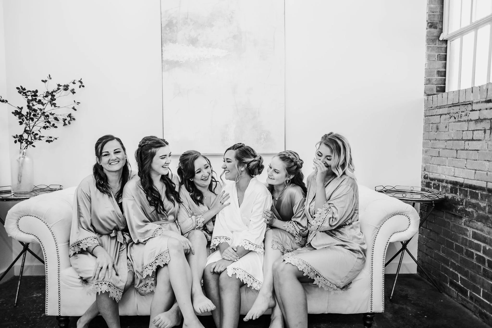 Six women sitting on a couch, laughing and smiling, dressed in matching robes, in a room with a brick wall and a large window.