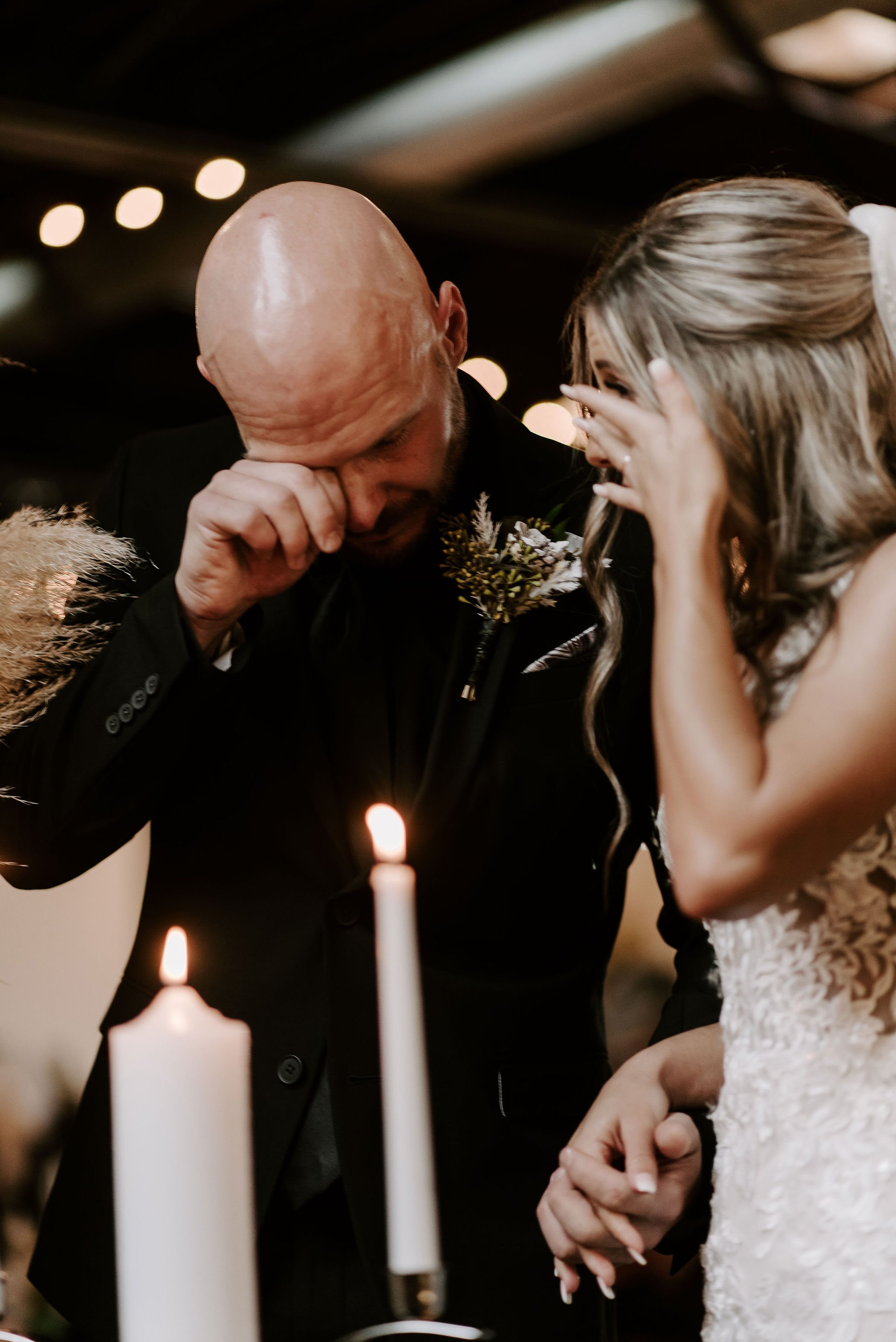 A man in a black suit with a boutonnière on wearing an emotional moment at a wedding, wiping tears as he holds a woman's hand, who is dressed in a lace wedding gown, with candles in the foreground and blurred lights in the background.