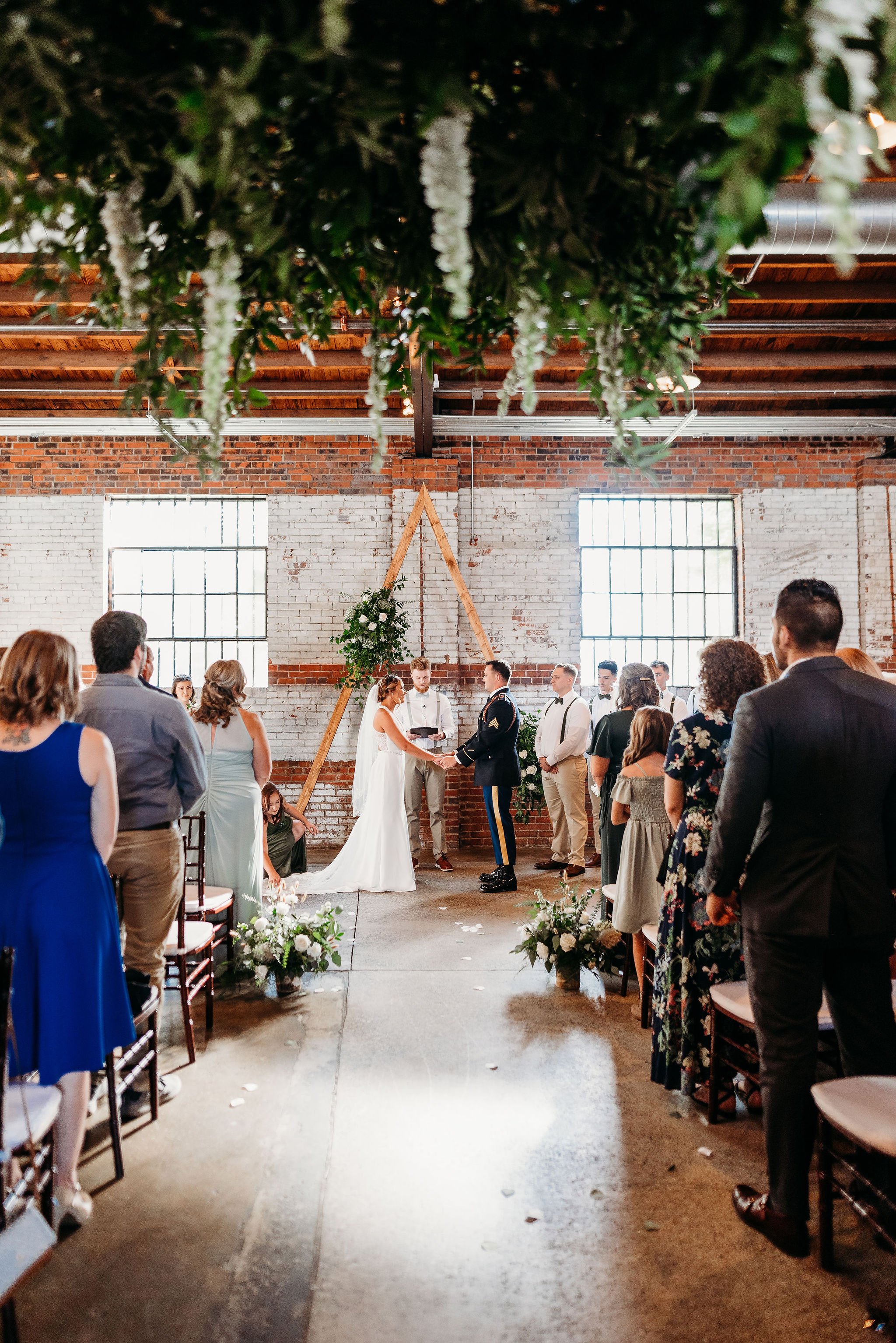 A wedding ceremony taking place in an industrial-style venue with exposed brick walls, large windows, and wooden ceiling beams. The bride and groom are exchanging vows at the altar decorated with greenery and flowers, while guests are seated and watc