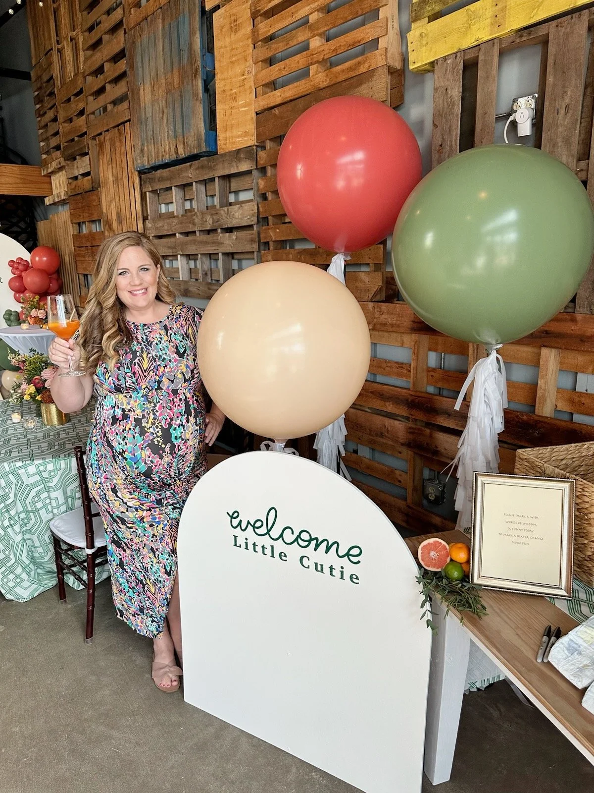 A woman in a colorful dress holding a glass of orange-colored drink, standing next to a sign that says 'welcome Little Cutie,' with three large balloons (red, green, beige) in the background, and a table decorated with flowers and fruit.
