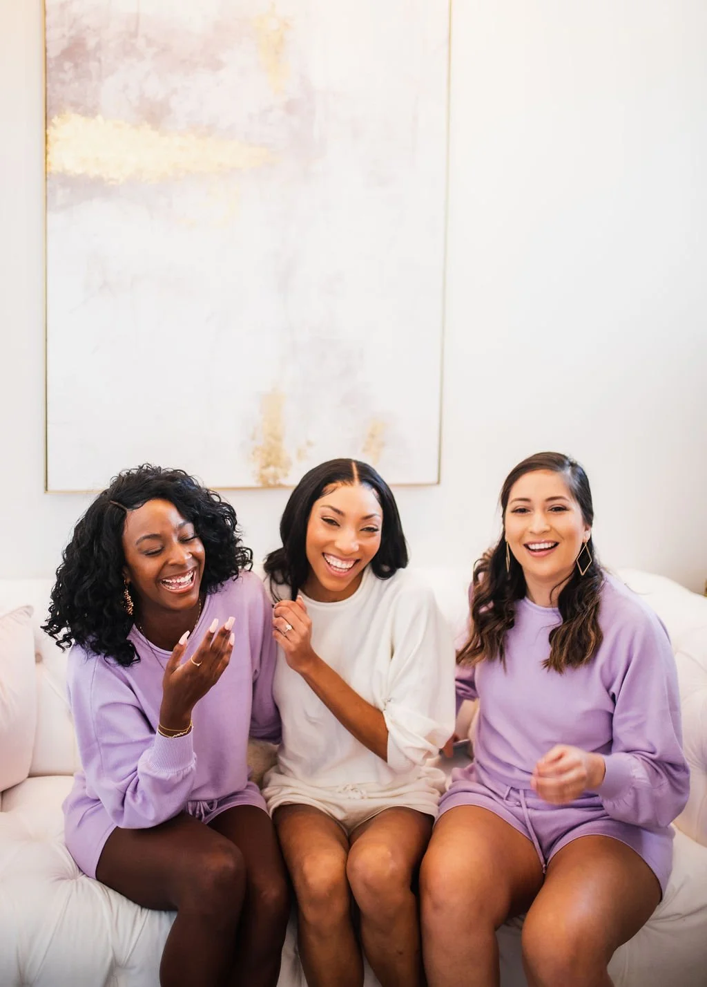Three women sitting on a white couch, smiling and laughing together. They are wearing casual clothing in pastel shades. The background features a light-colored wall with a large abstract painting.