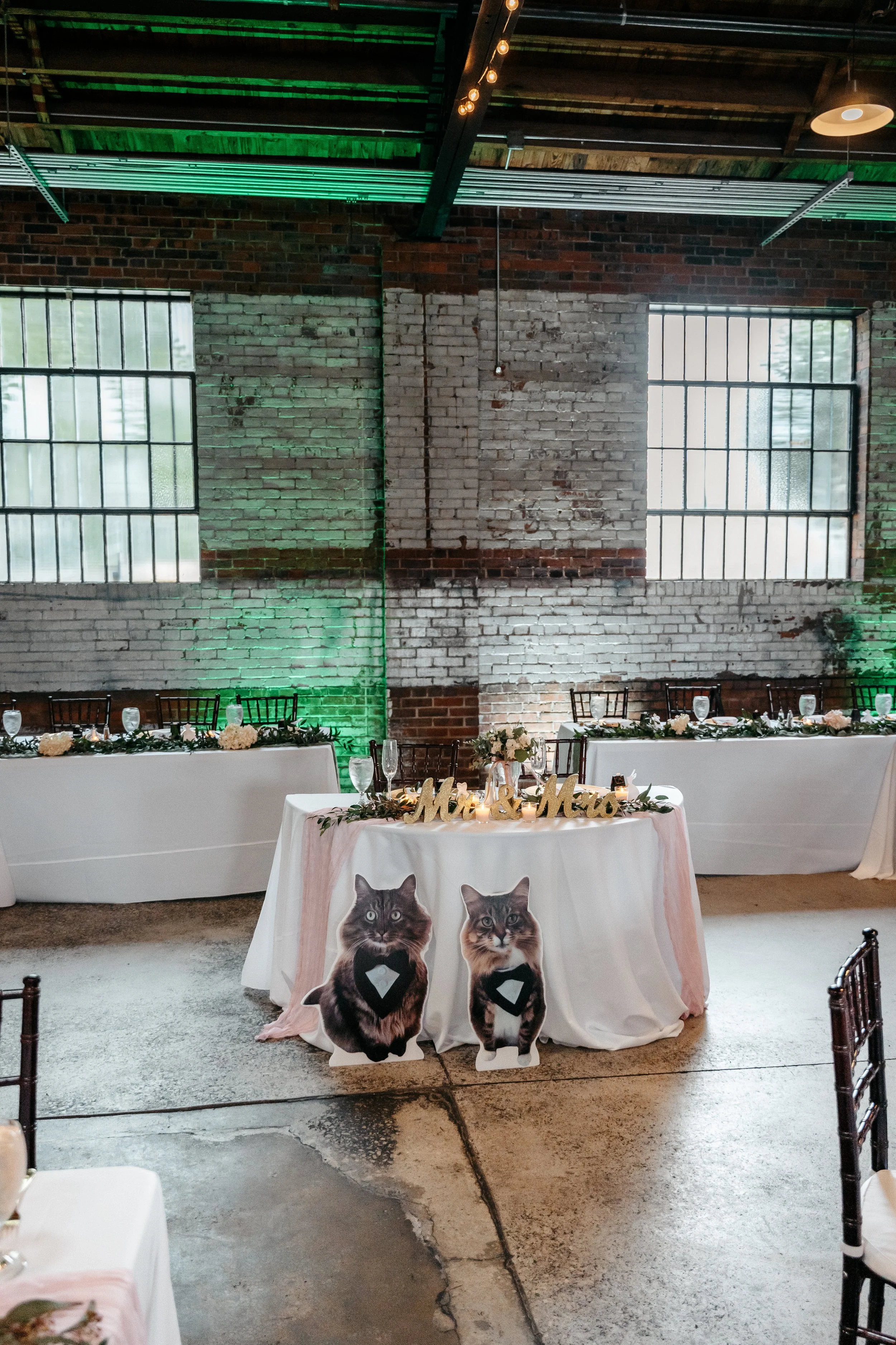Wedding reception table decor with two cat cutouts dressed as grooms, 'Mr. & Mrs.' sign, floral arrangements, and place settings in an industrial-style venue with exposed brick walls and large windows.