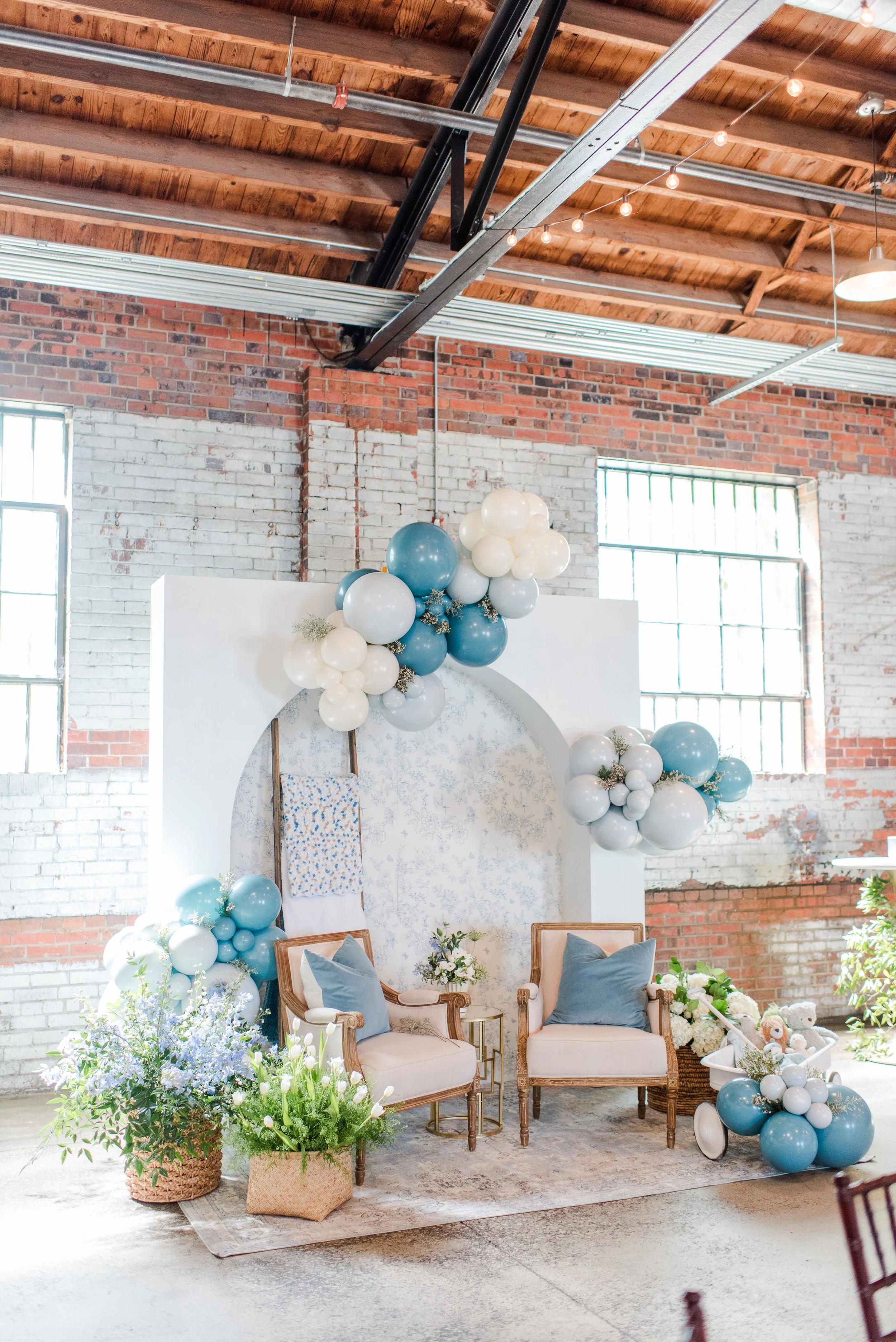 Decorative setup with two vintage armchairs with blue pillows, surrounded by blue and white balloons, flowers, and teddy bears in a rustic, industrial-style space with exposed brick walls and wood ceiling.