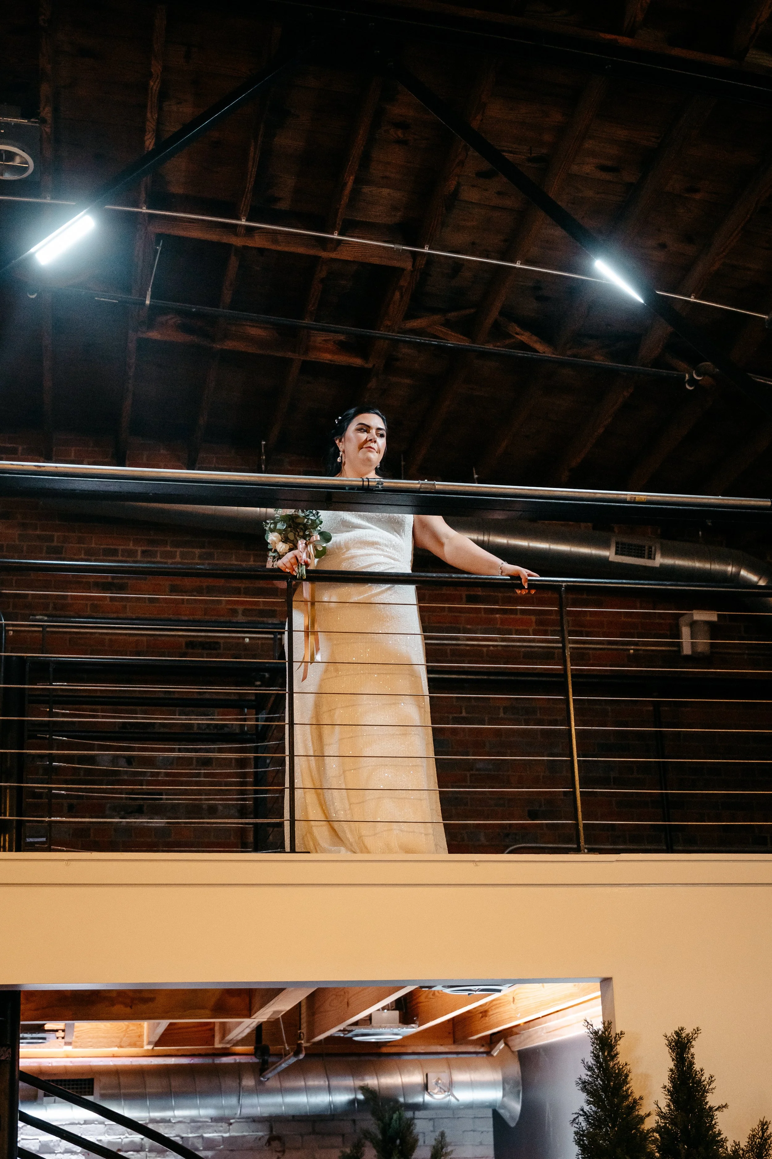 A woman in a white dress holding a bouquet standing on a balcony with a brick wall and wooden ceiling in the background.