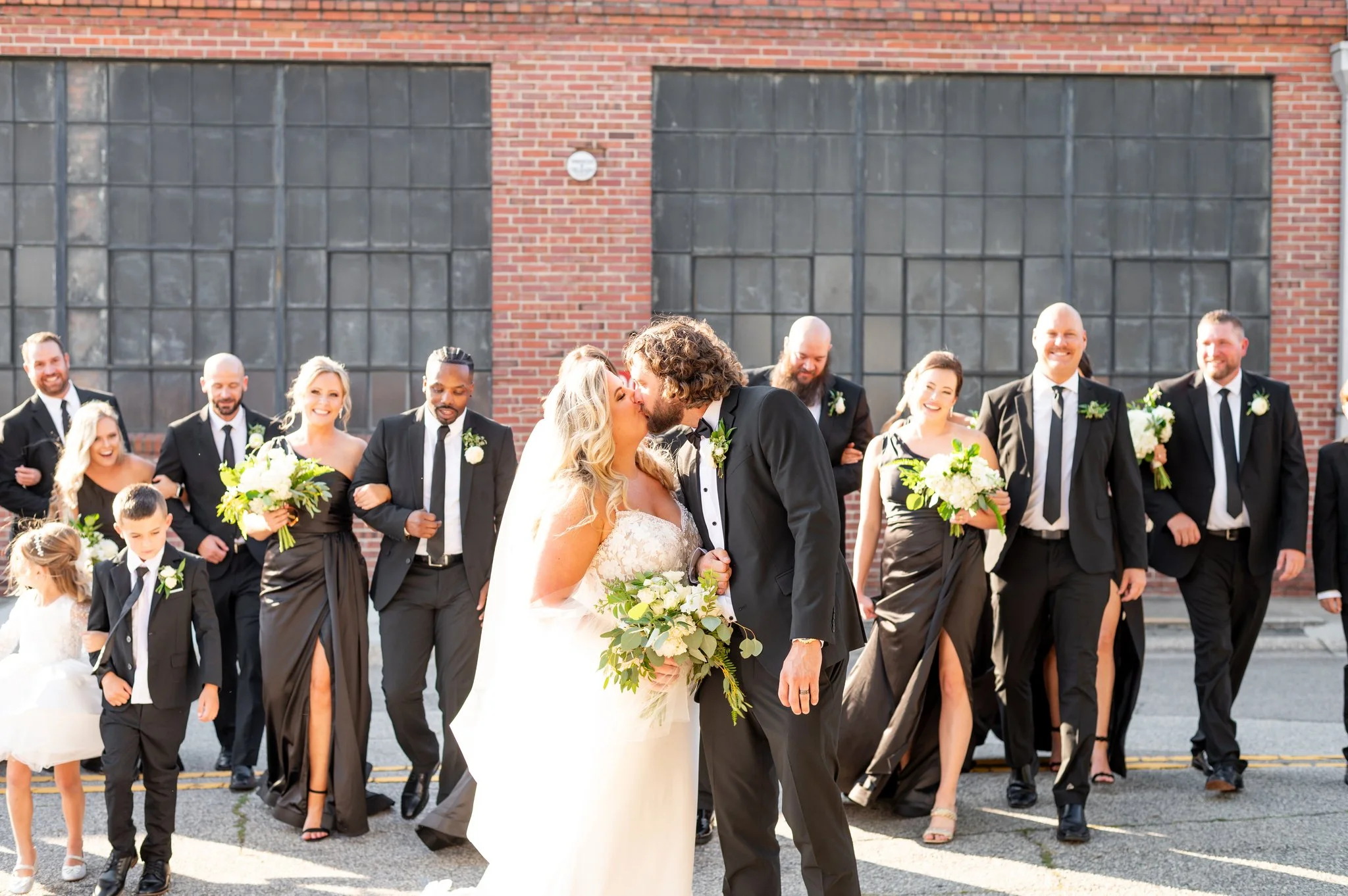 Wedding couple kissing in front of bridesmaids and groomsmen on city street, with brick building background.