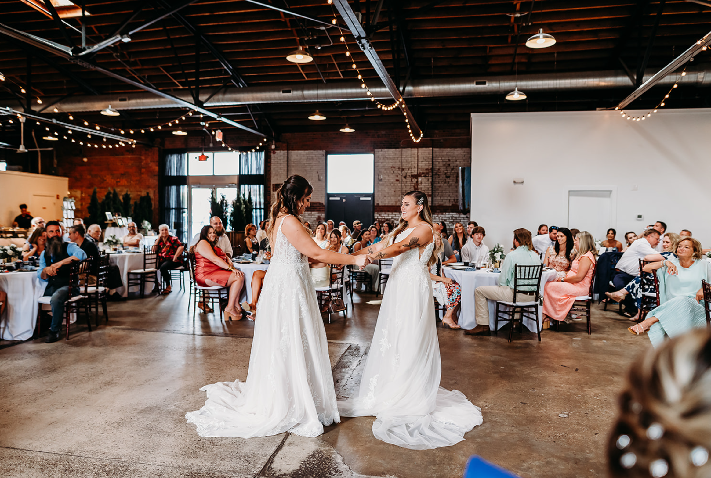 Two brides holding hands and smiling at each other during their wedding ceremony in a spacious, industrial-style venue with exposed brick walls and string lights, while guests sit at decorated tables around them.