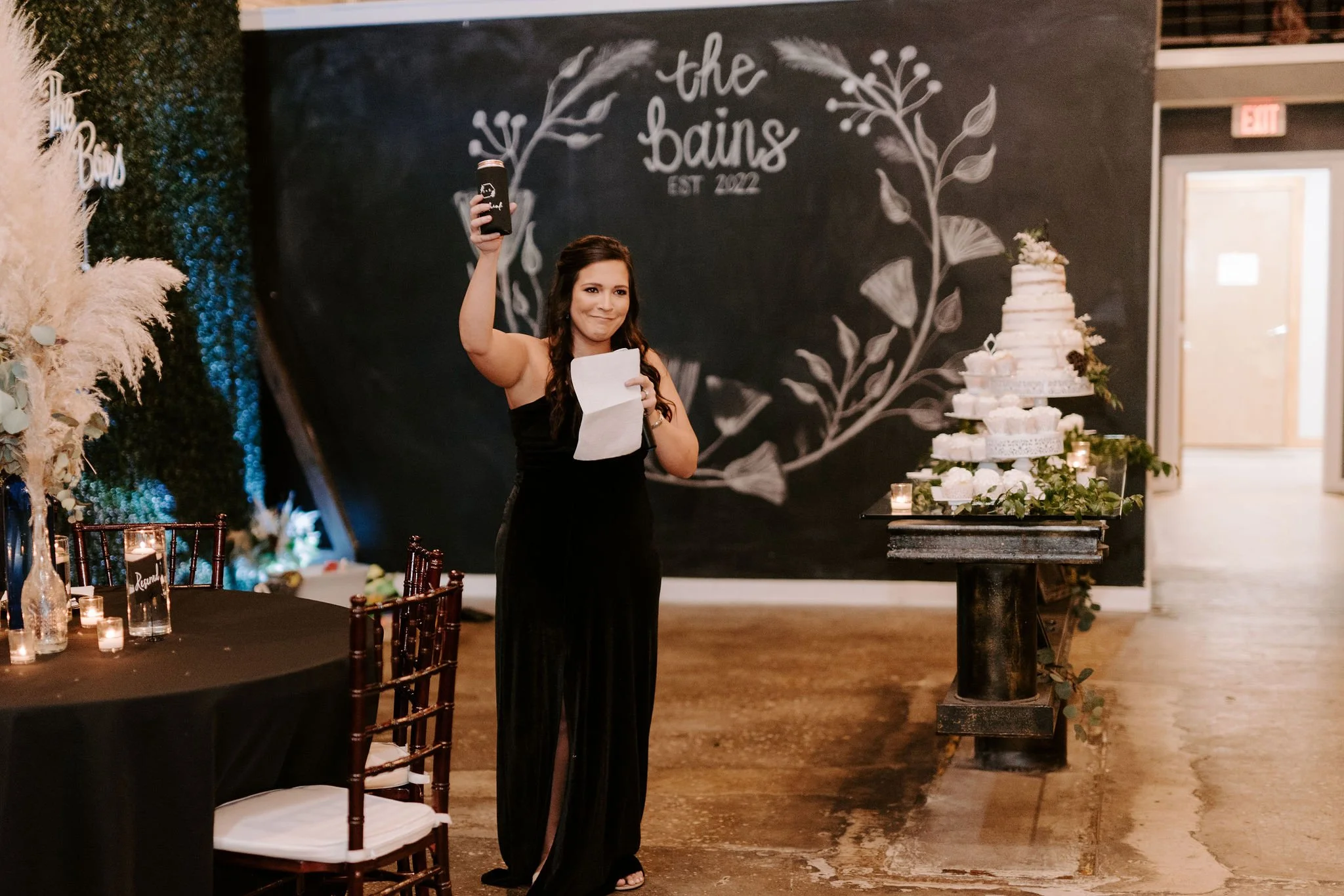 A woman in a black dress holding a can and a piece of paper while standing near a wedding cake at an indoor reception with a black chalkboard wall and floral decorations.